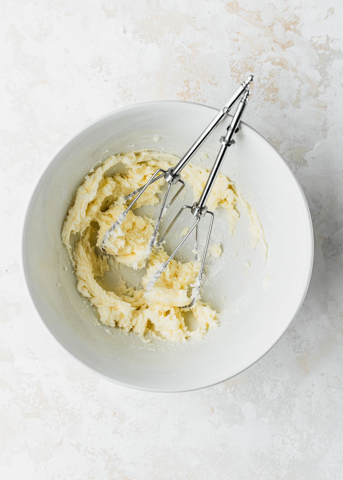 A white bowl of mixed butter, powdered sugar, and beaters on a white counter.