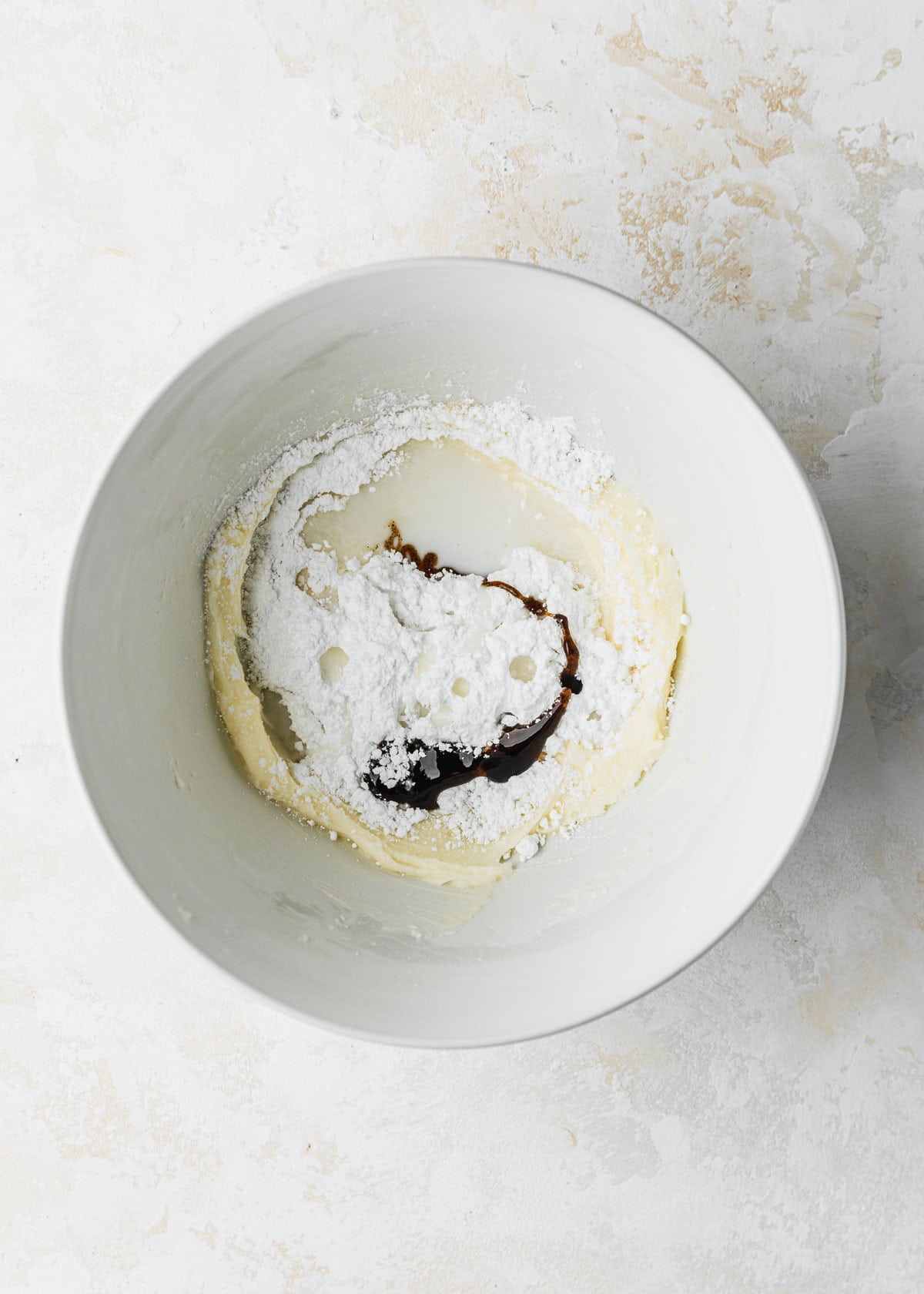 A white bowl of butter, powdered sugar, milk, and vanilla bean paste on a white table.