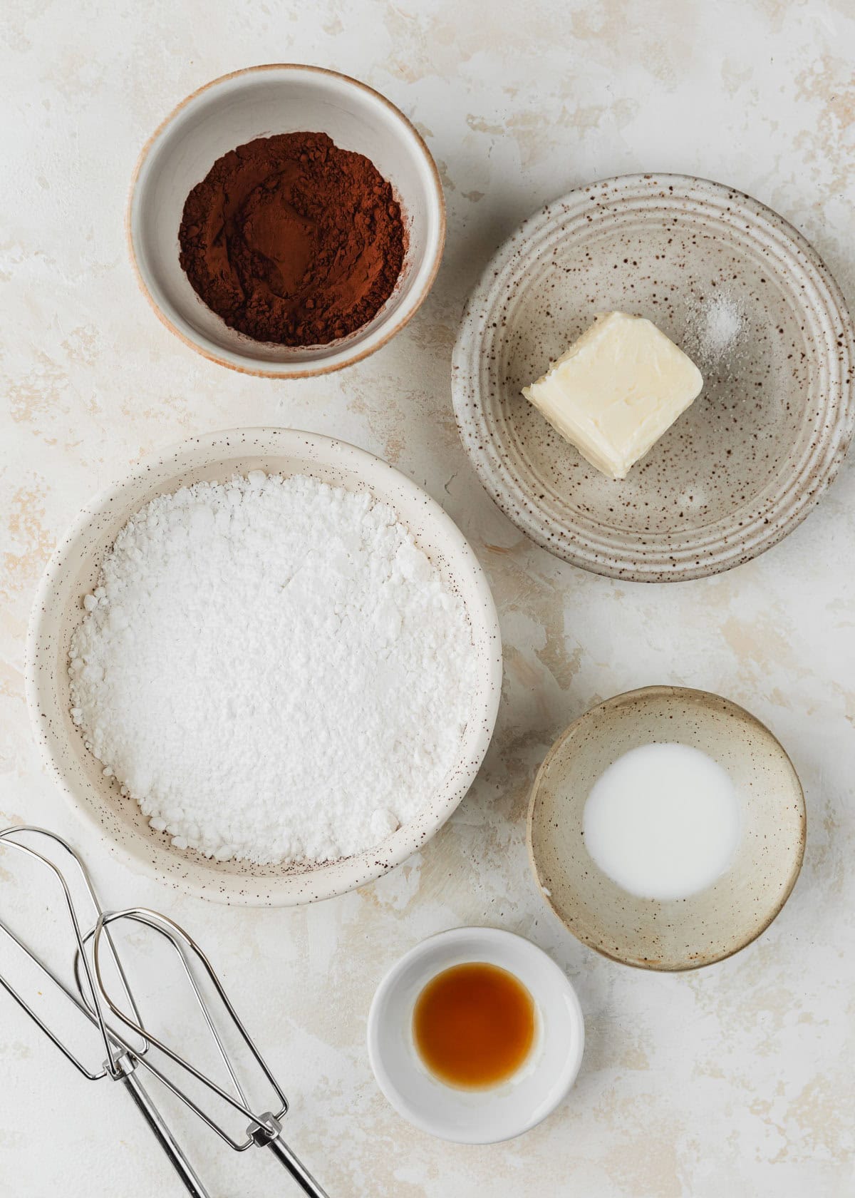 White bowls of powdered sugar, butter, cocoa powder, milk, and vanilla extract on a tan counter next to beaters.