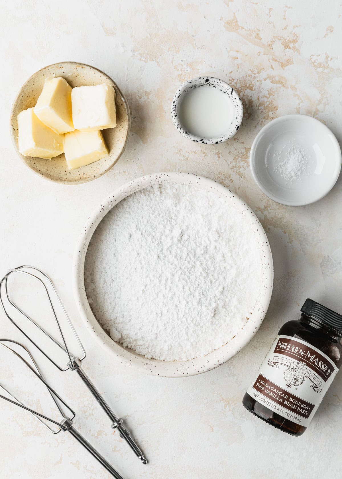 White bowls of butter, salt, powdered sugar, and milk on a beige table next to beaters and a jar of vanilla bean paste.
