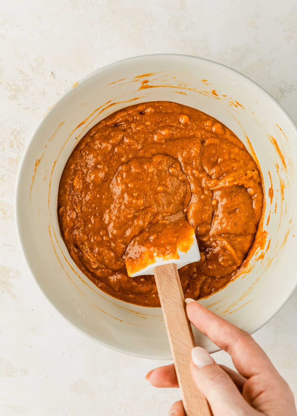 A hand using a rubber spatula to mix batter for small batch pumpkin muffins in a white bowl.