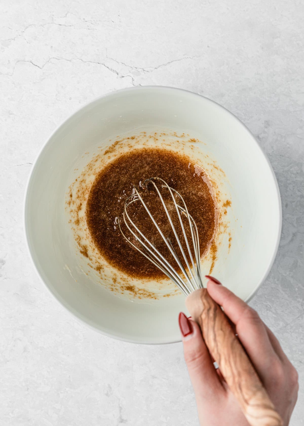 A hand using a whisk to mix butter and sugar in a white bowl.