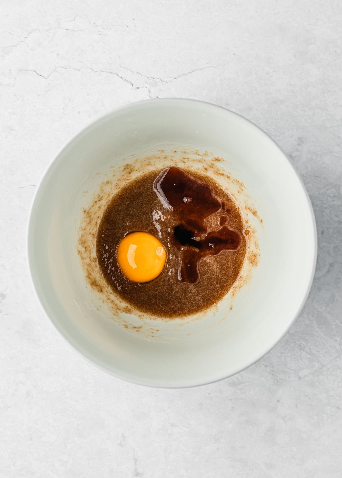 A white bowl of butter, brown sugar, an egg yolk, and vanilla on a white counter.