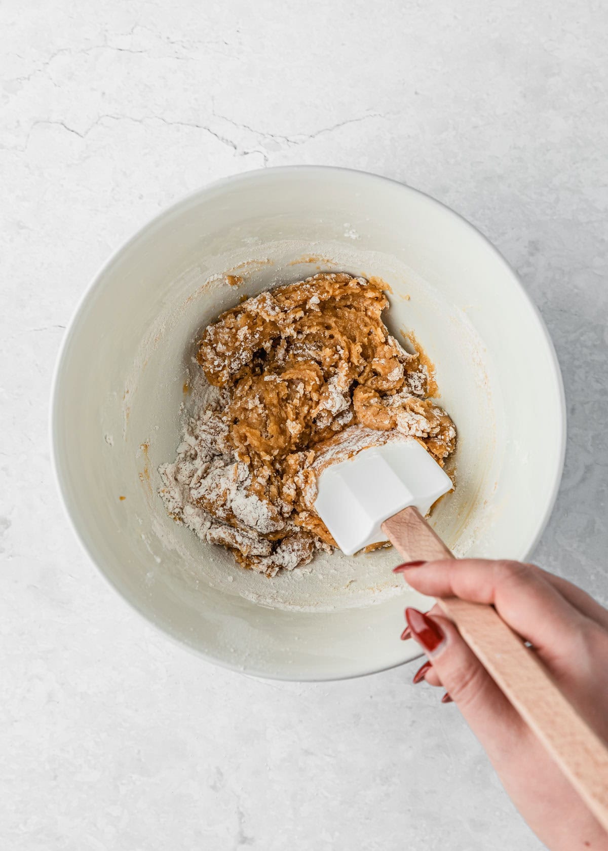 A hand using a rubber spatula to mix dough for small batch chocolate chip cookie bars in a white bowl.