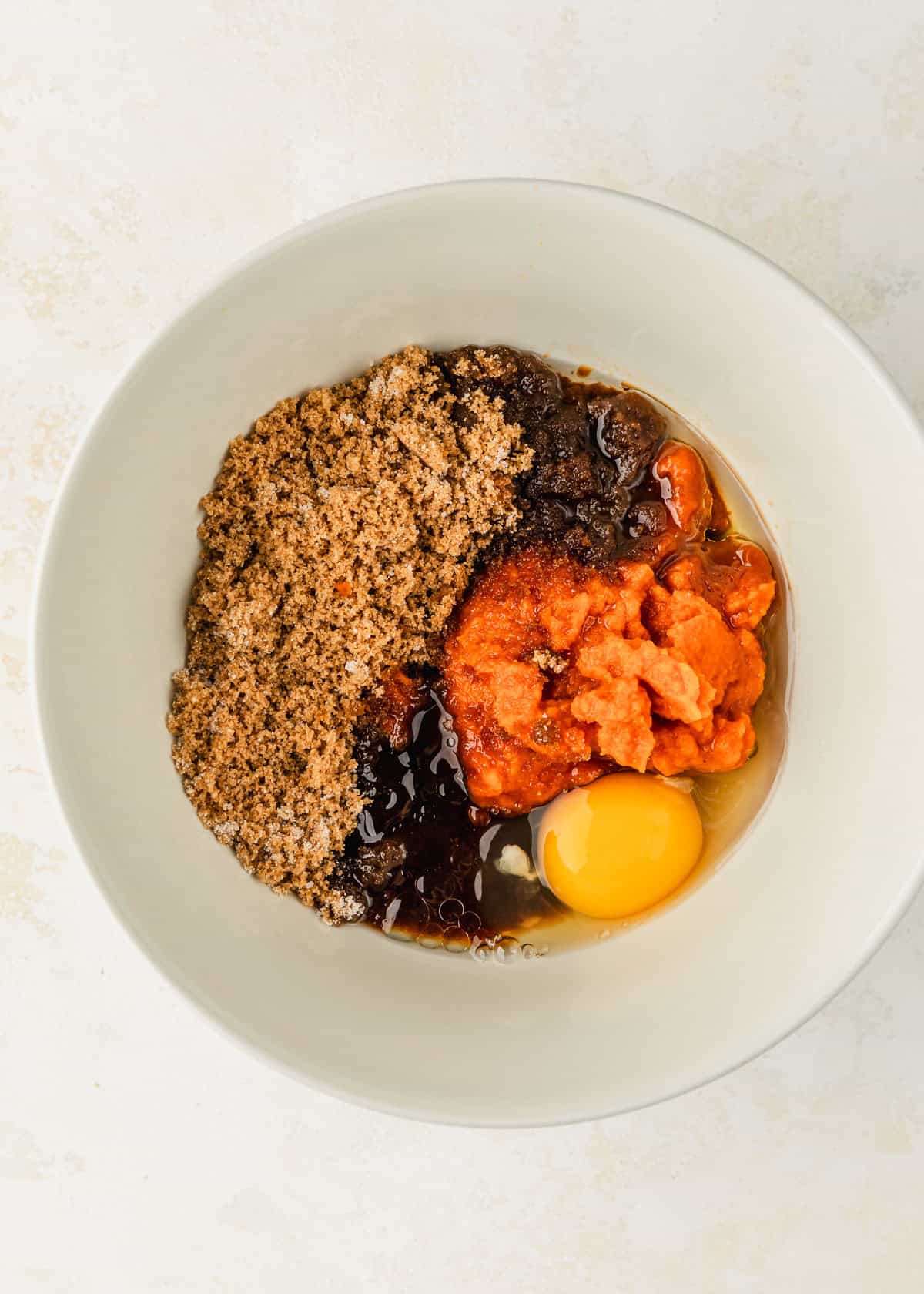 A white bowl of oil, pumpkin, an egg, and sugar on a tan counter.