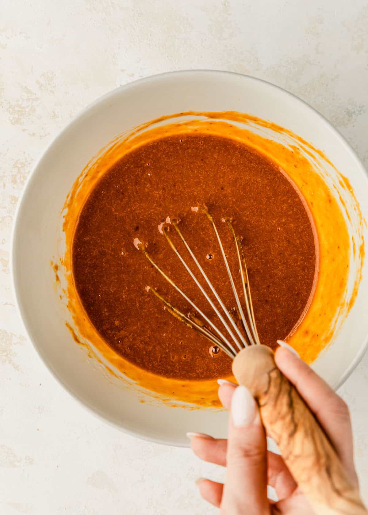 A hand whisking pumpkin filling for small batch pumpkin muffins in a white bowl on a beige table.