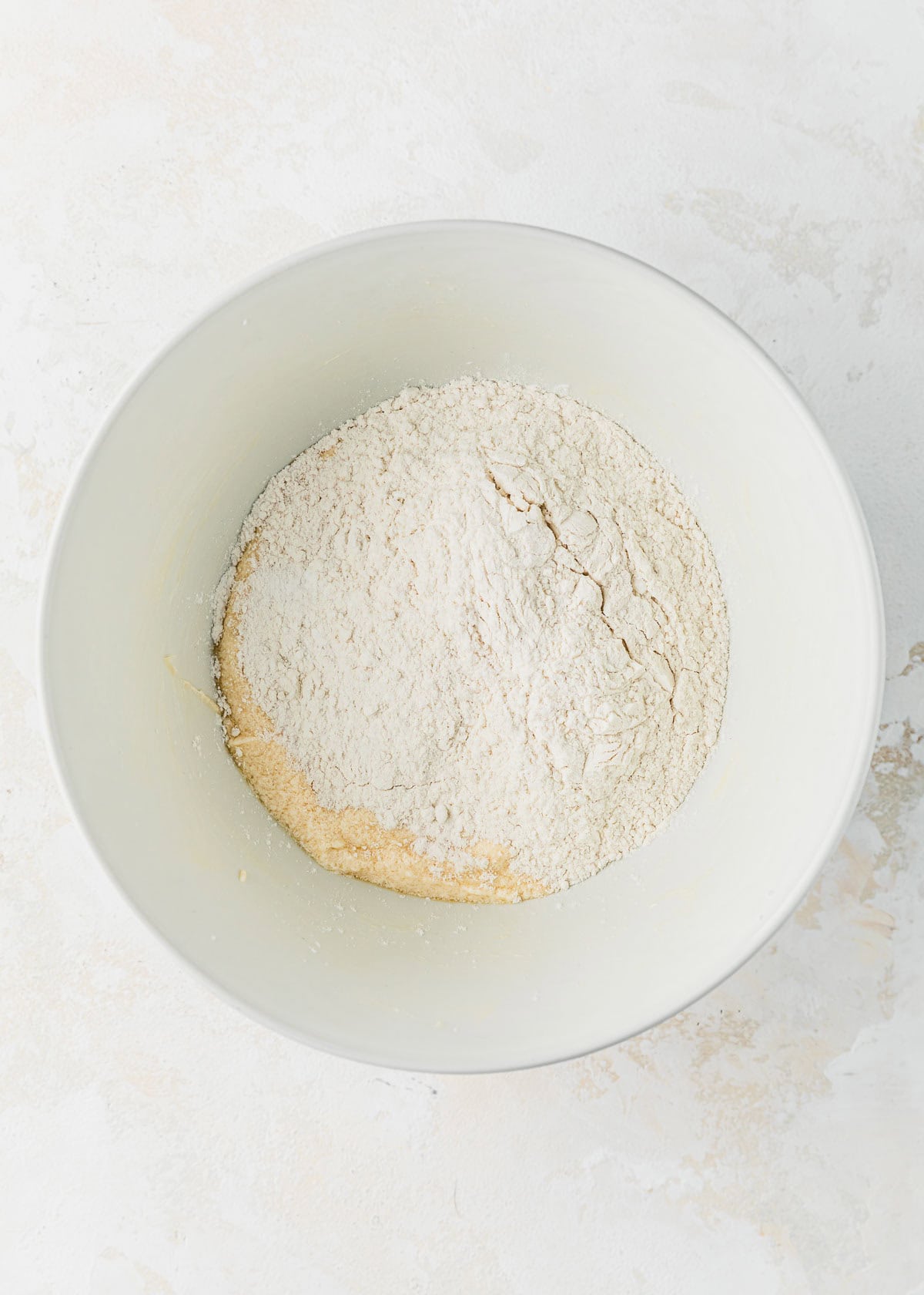 A white bowl of whipped butter, sugar, and eggs topped with flour on a white counter.