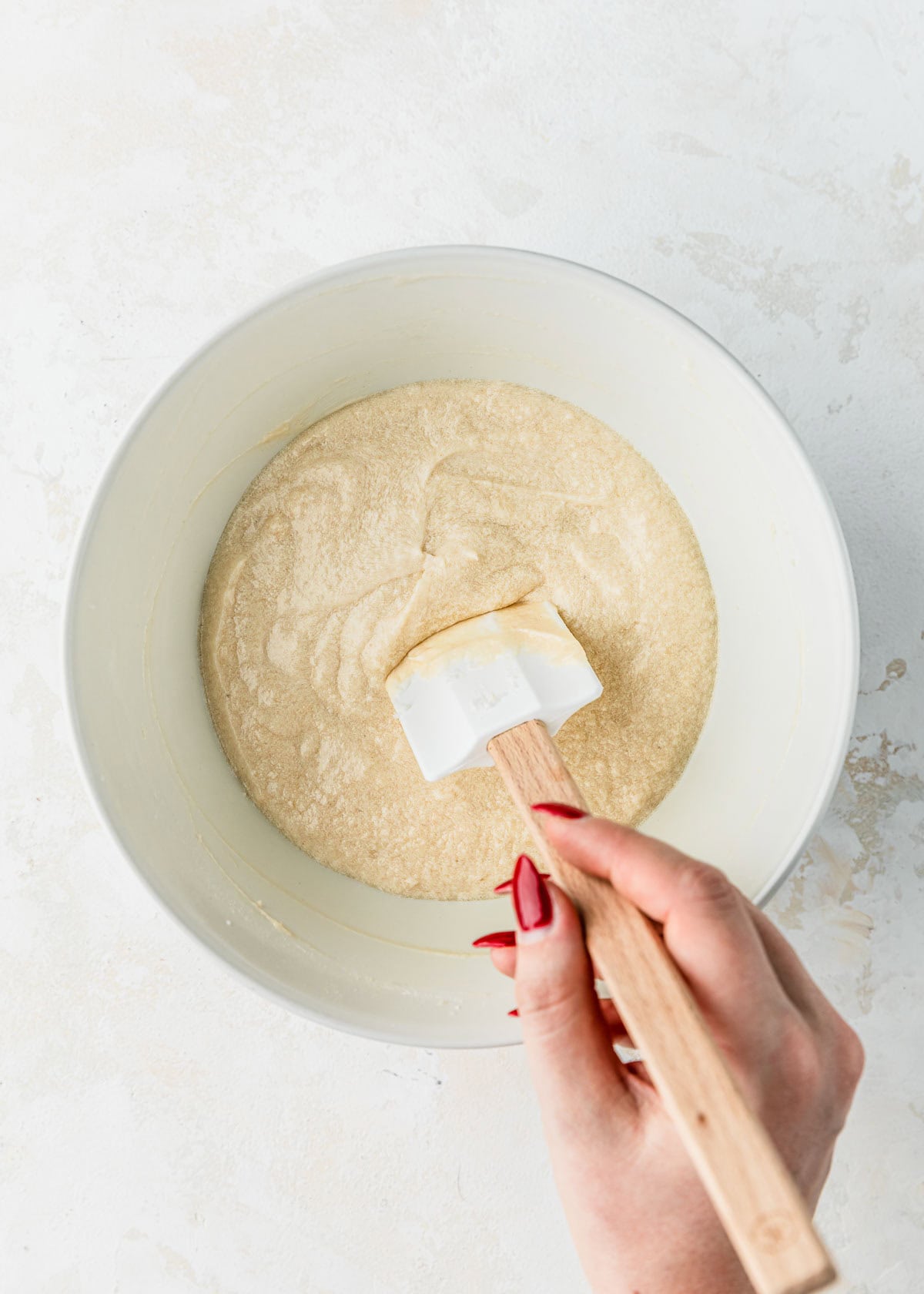 A hand using a rubber spatula to mix a white bowl of batter for small batch vanilla cupcakes.