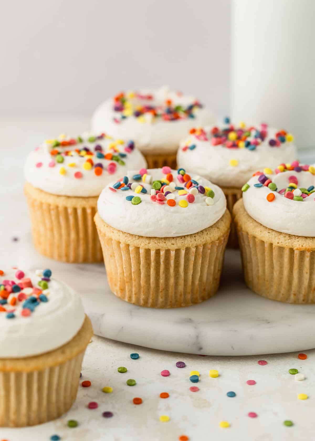 A white tray of small batch vanilla cupcakes with rainbow sprinkles on a tan background next to more sprinkles and a glass of milk.