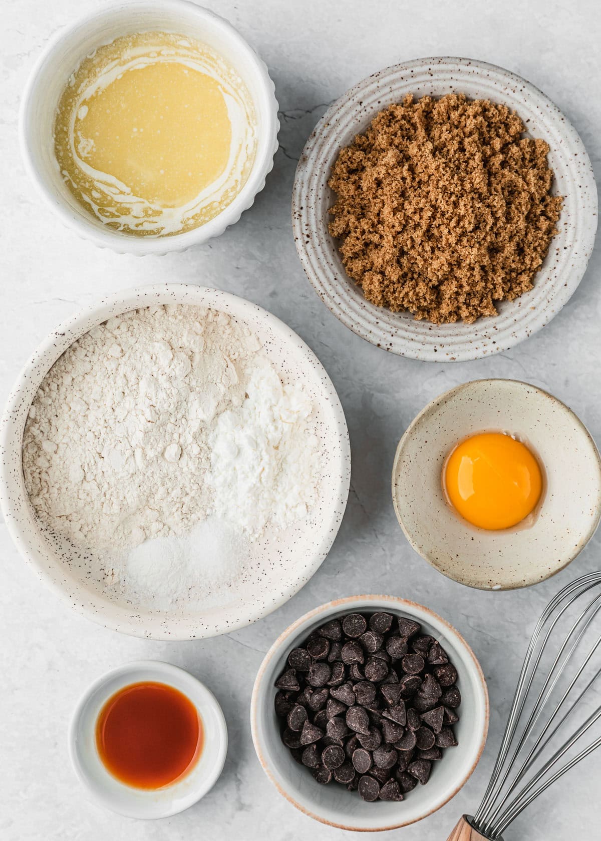 White bowls of flour, cornstarch, salt, baking powder, butter, brown sugar, an egg yolk, vanilla, and chocolate chips on a white counter next to a whisk.
