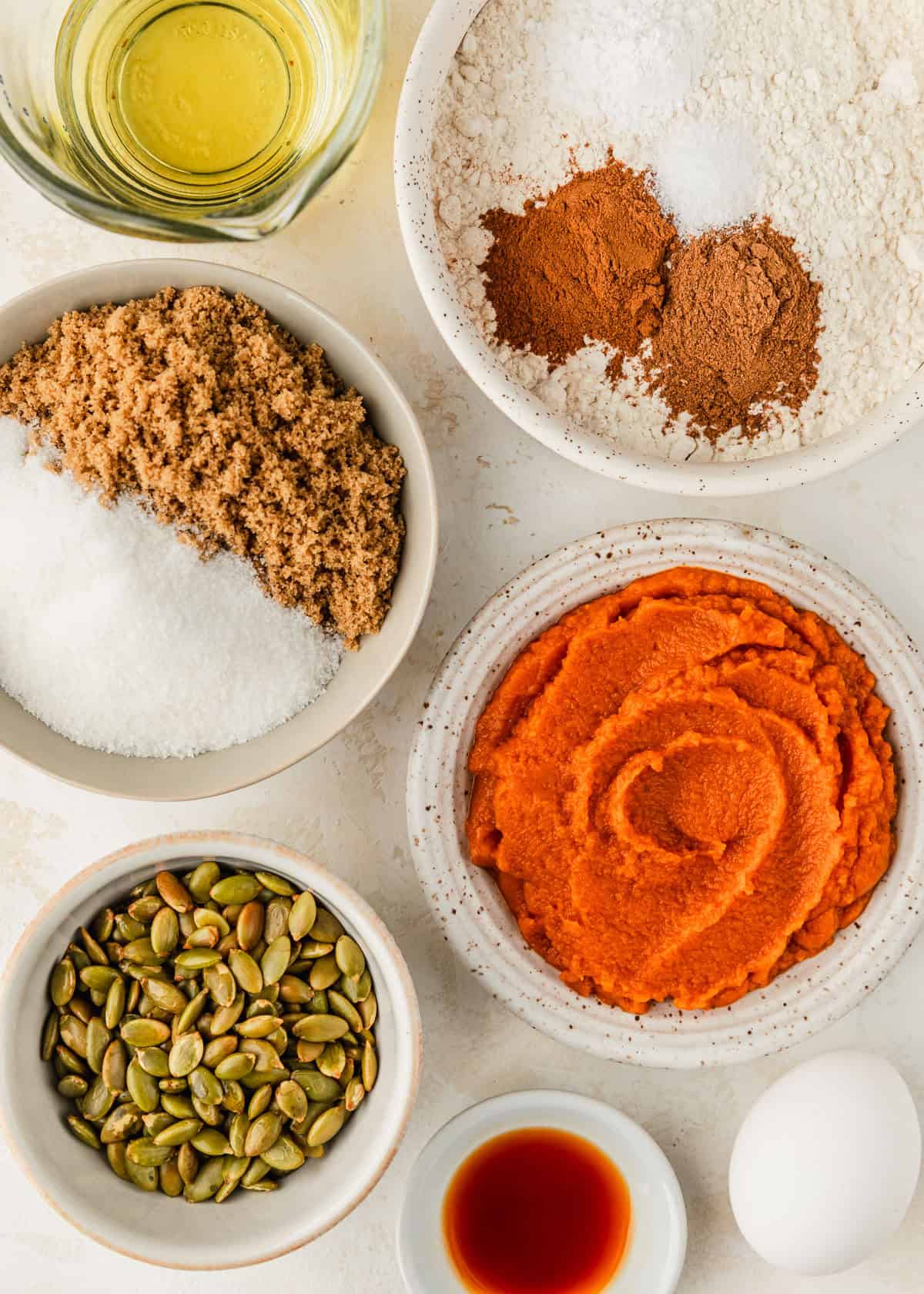 White bowls of flour, spices, pumpkin, brown and white sugar, pumpkin seeds, and vanilla on a tan counter next to an egg and glass of oil.