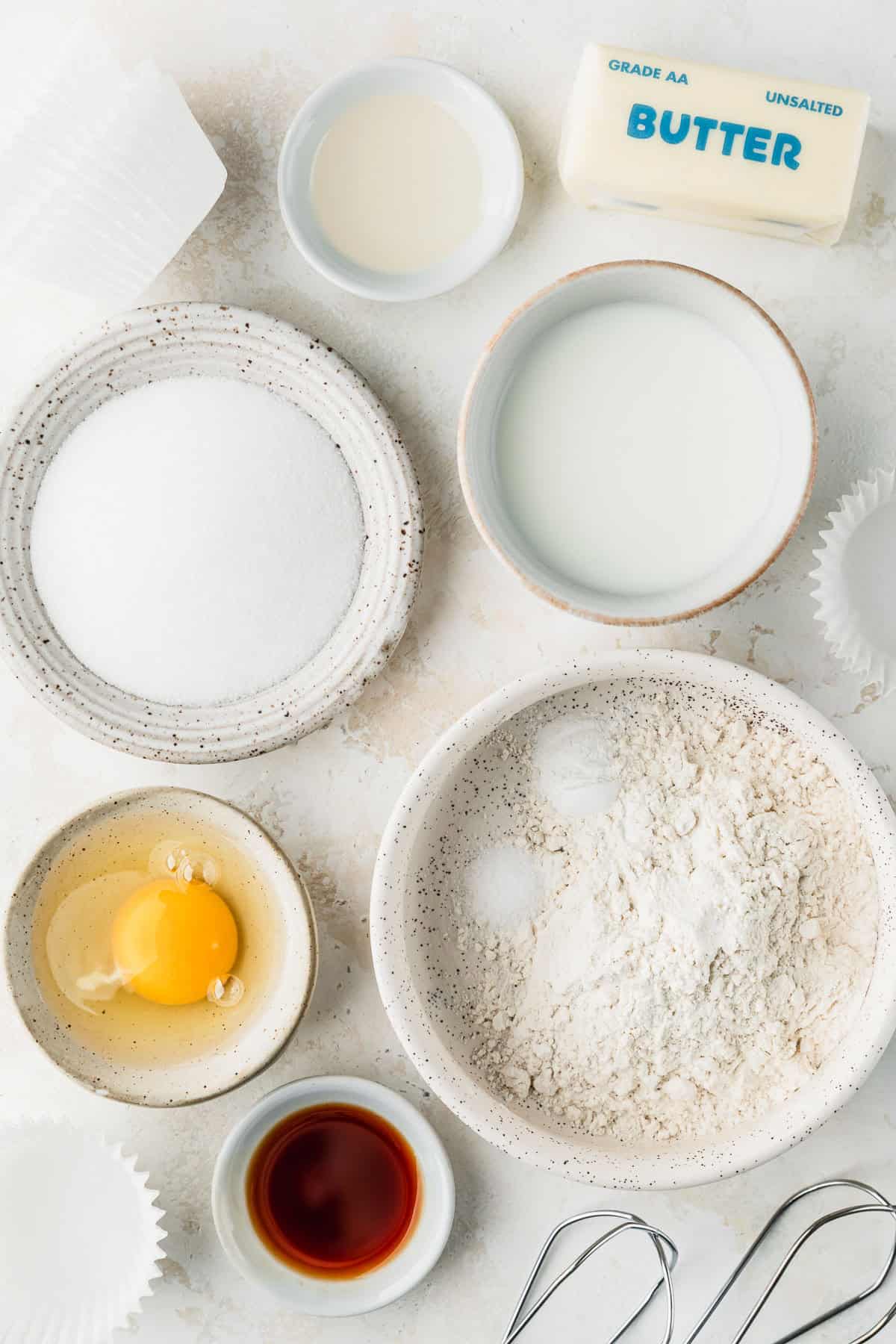 White bowls of flour, milk, sugar, oil, an egg, and vanilla next to a stick of butter on a light tan counter.
