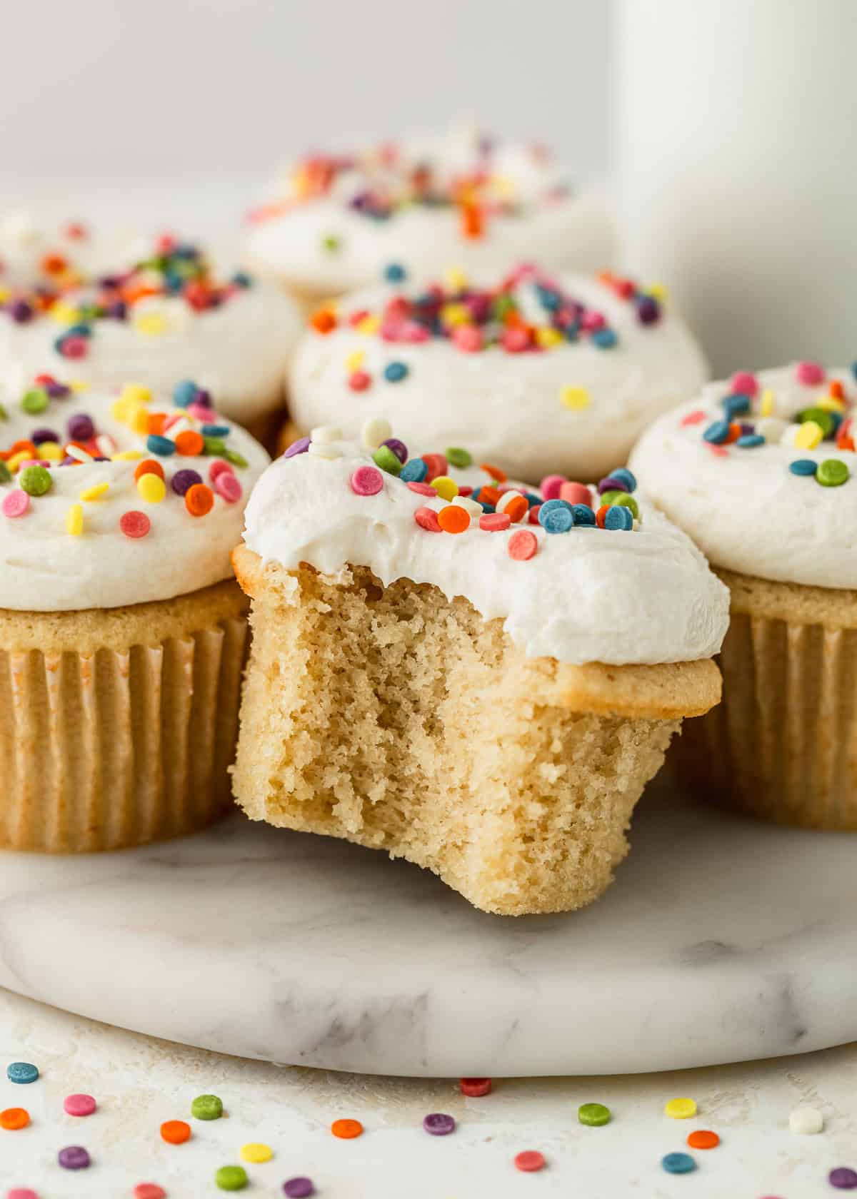 Rows of small batch vanilla cupcakes with rainbow sprinkles on a white board next to a glass of milk with a cream-colored background.