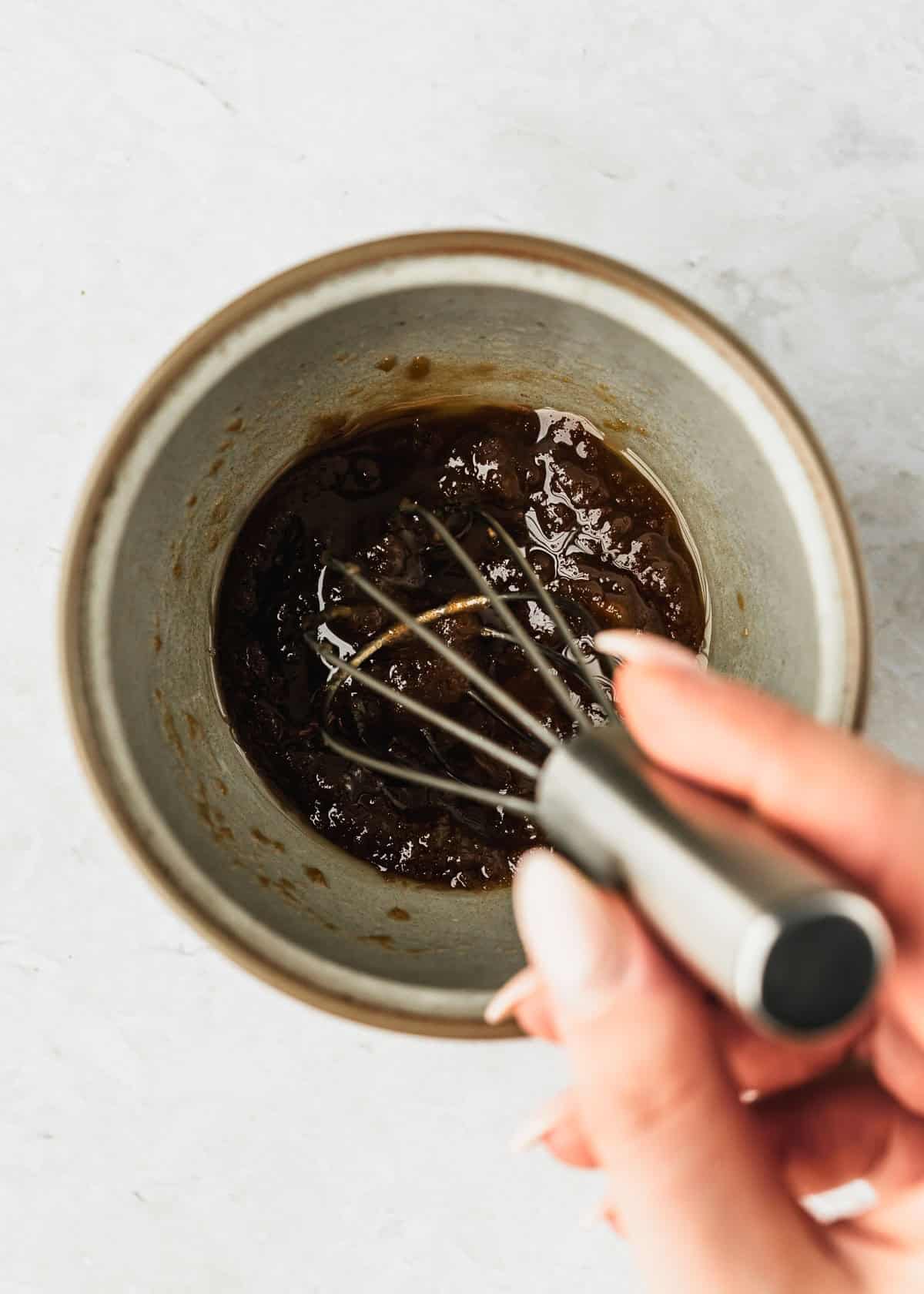 A hand using a whisk to mix butter and sugar in a grey mug.