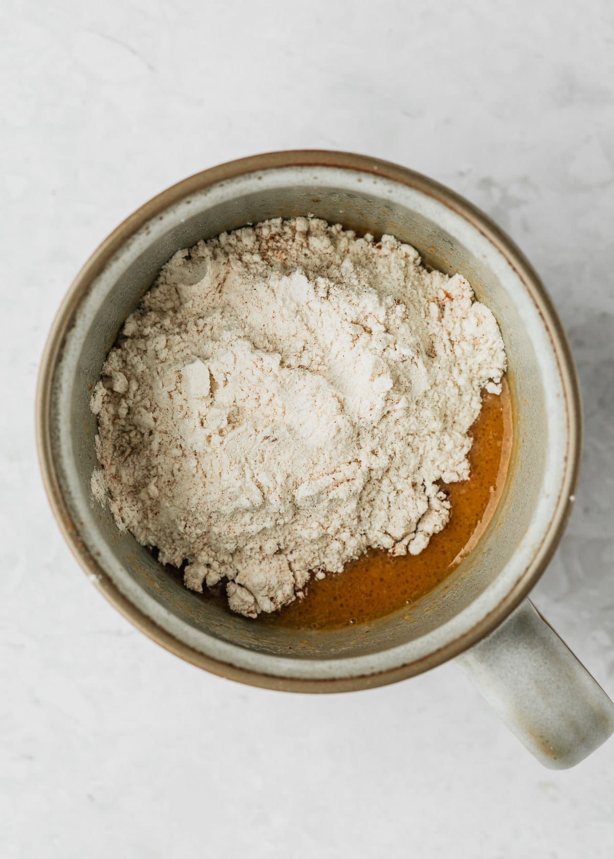 A grey mug with pumpkin, flour, and spices on a white counter.