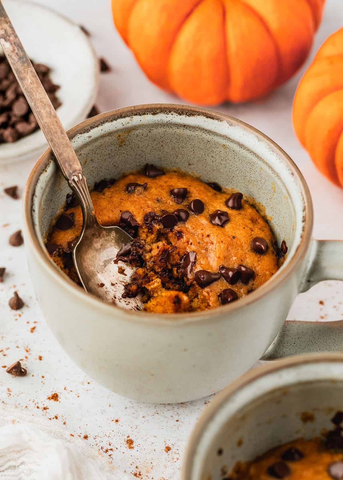 A pumpkin mug cake on a white counter next to pumpkins, a white napkin, and a white bowl of chocolate chips.