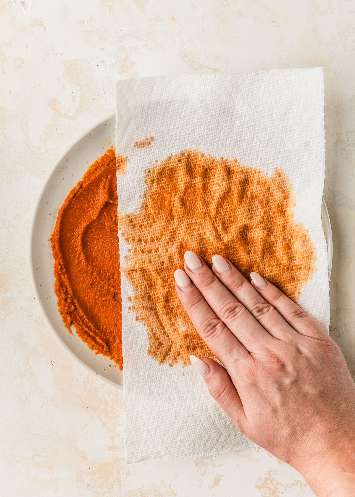 A hand pressing a paper towel onto a white plate of pumpkin puree.