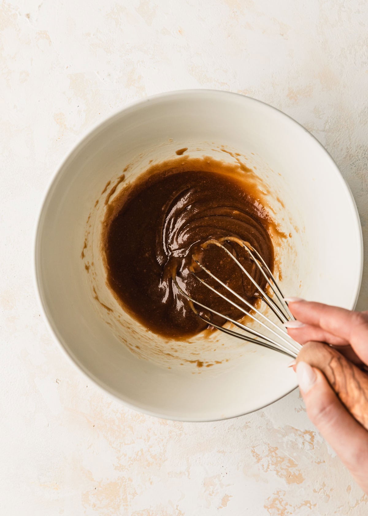 A hand using a whisk to mix melted butter and brown sugar in a white bowl.