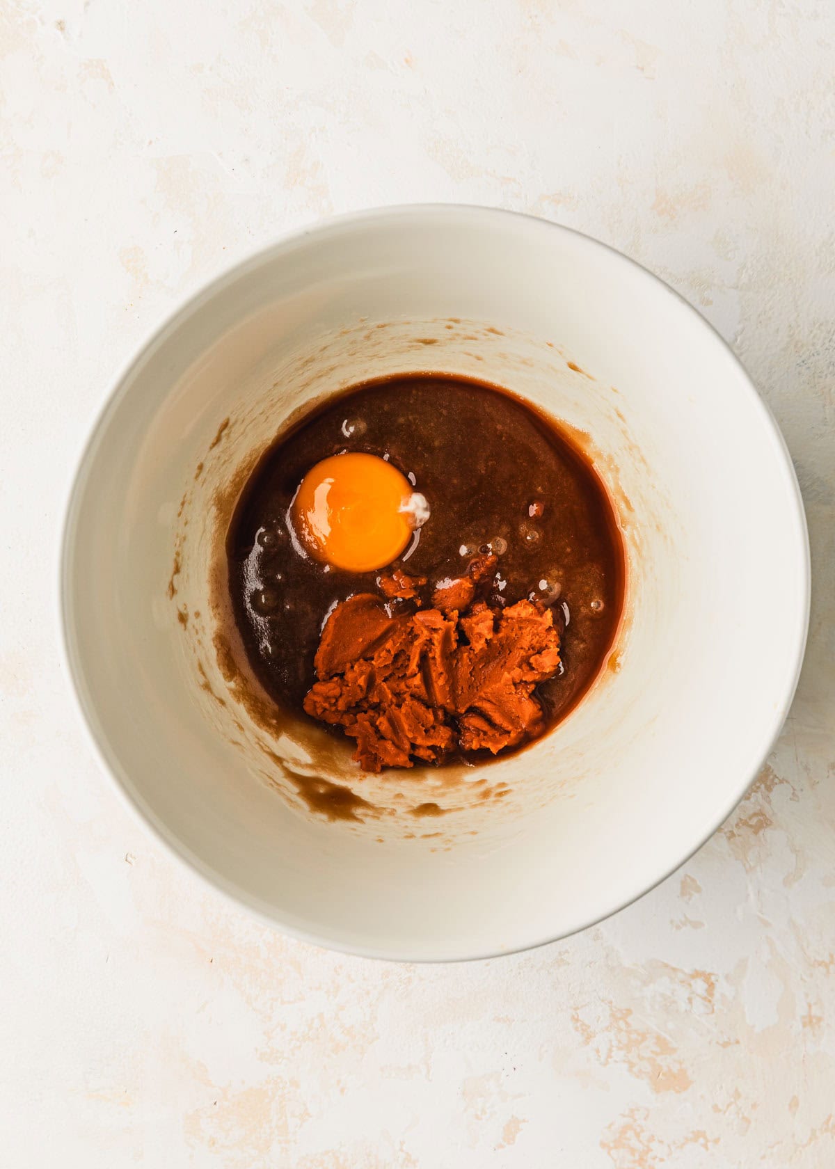 A white bowl of butter, sugar, an egg yolk, vanilla, and pumpkin puree for small batch pumpkin cookies on a tan counter.