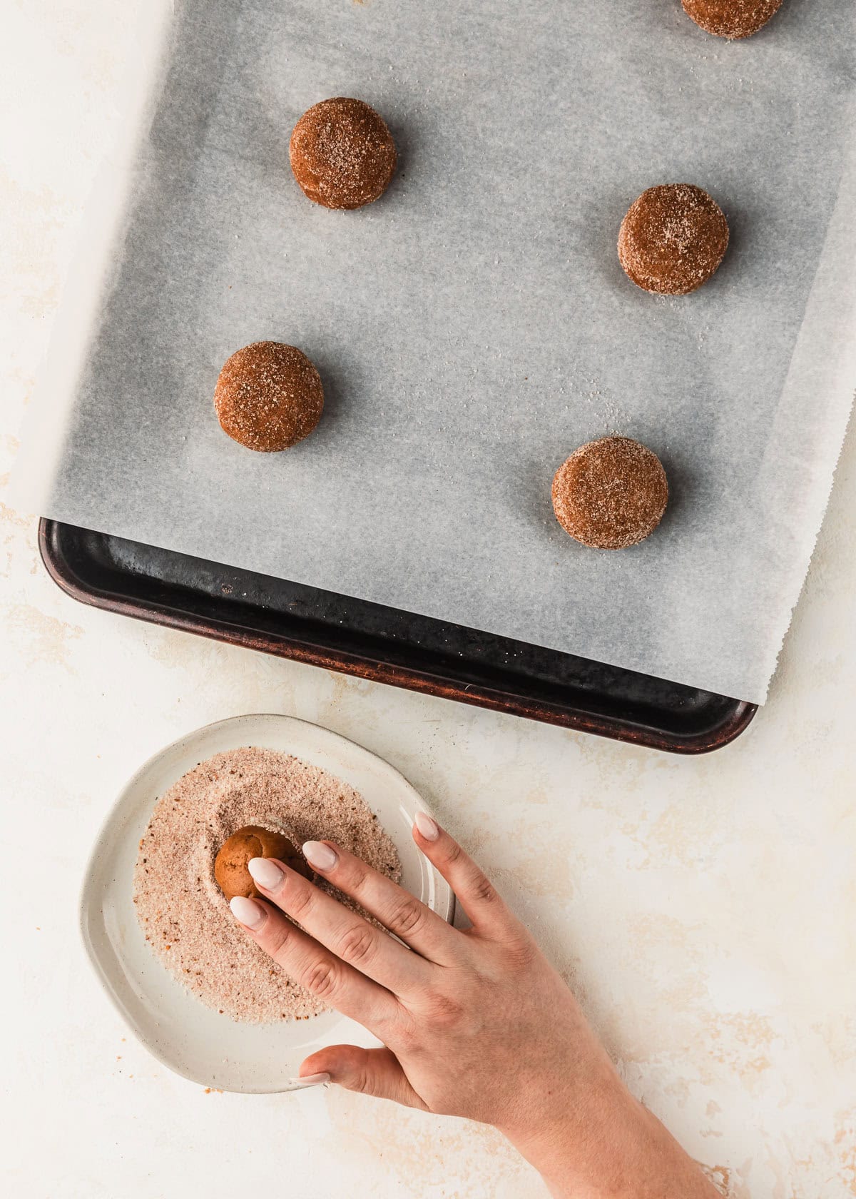 A hand rolling cookie dough in cinnamon sugar next to a sheet pan of small batch pumpkin cookies.