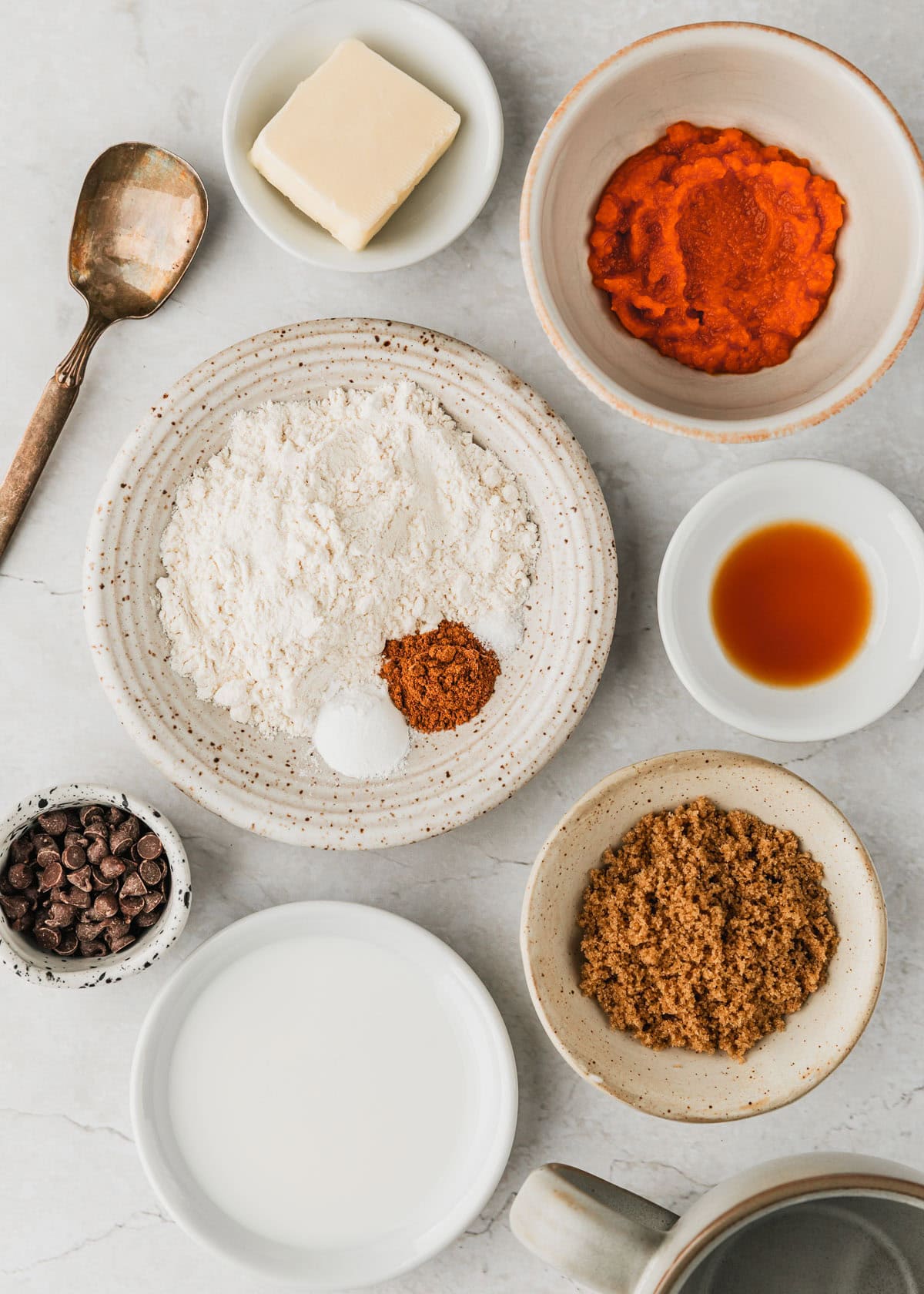 White bowls of butter, pumpkin puree, vanilla, flour, spices, brown sugar, milk, and chocolate chips next to a grey mug on a white marble counter.