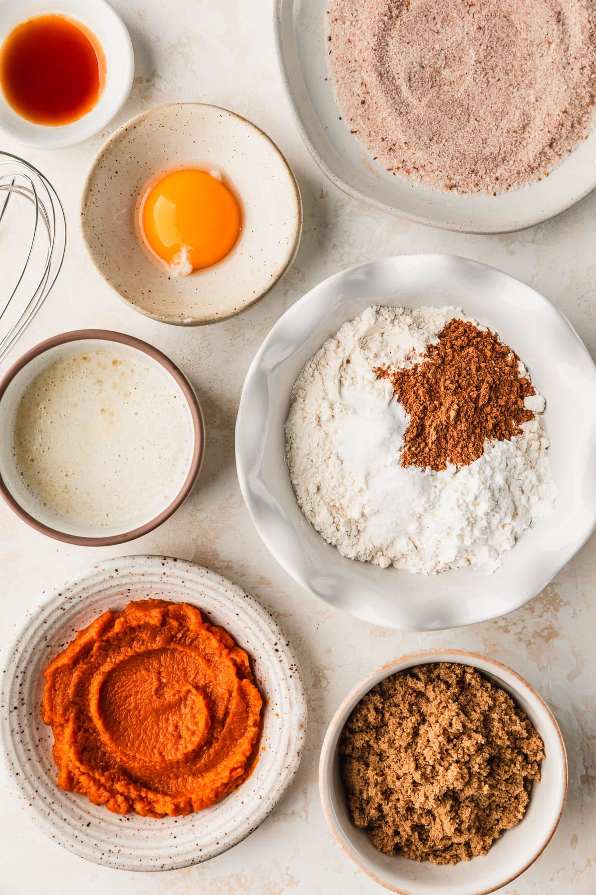 White bowls of pumpkin puree, brown sugar, butter, flour, spices, an egg yolk, vanilla, and cinnamon sugar on a tan counter.