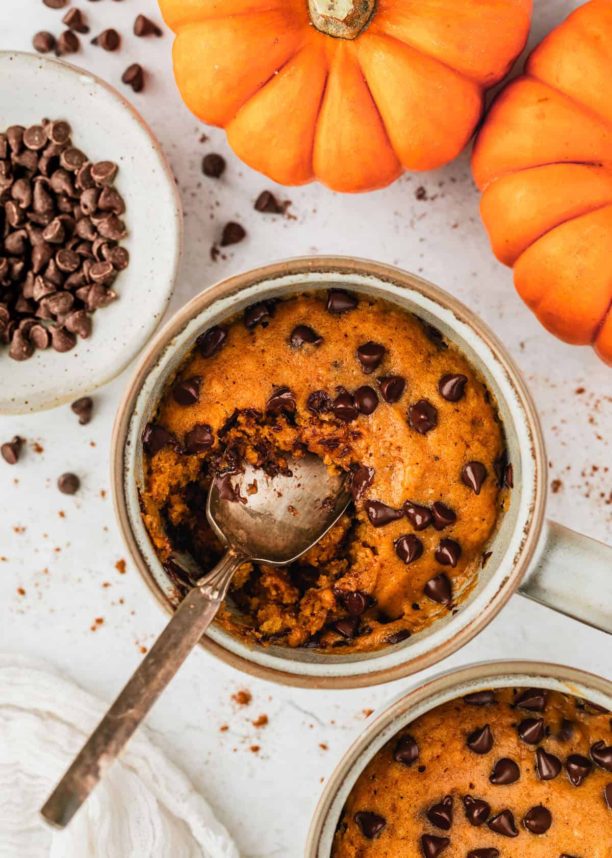 A spoon digging into a pumpkin mug cake on a white counter next to mini pumpkins, another cake, and a white bowl of chocolate chips.