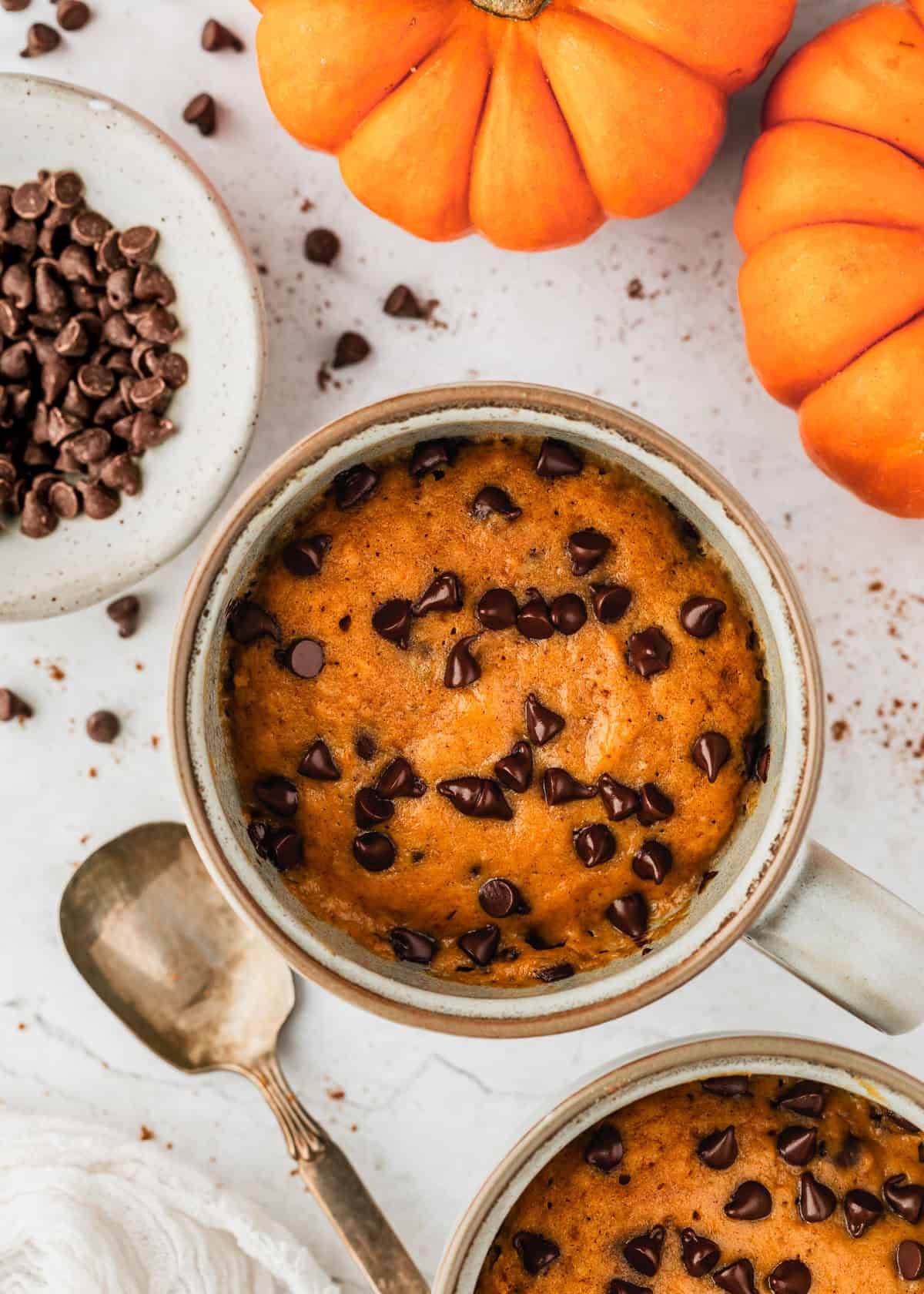 Two pumpkin mug cakes on a white counter next to pumpkins, a spoon, and a white bowl of chocolate chips.