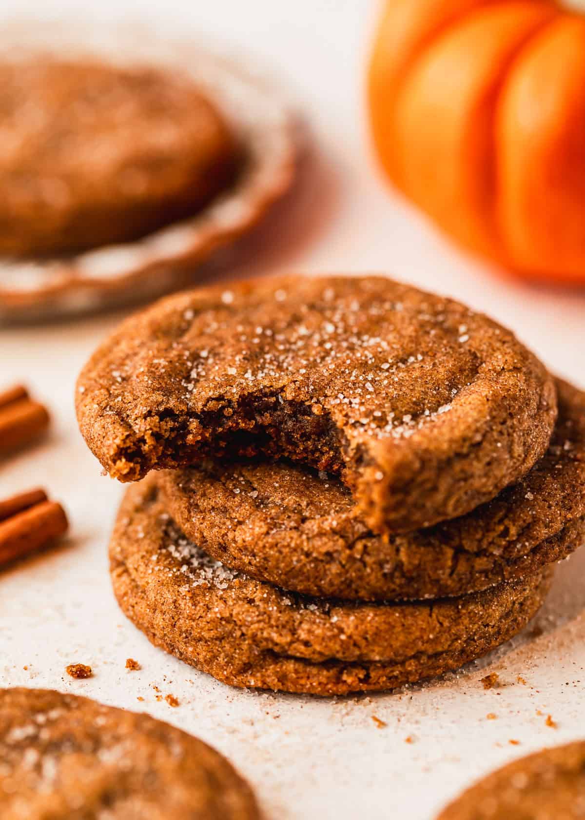 A stack of small batch pumpkin cookies next to a pumpkin, more cookies, a white plate, and cinnamon sticks with a tan background.