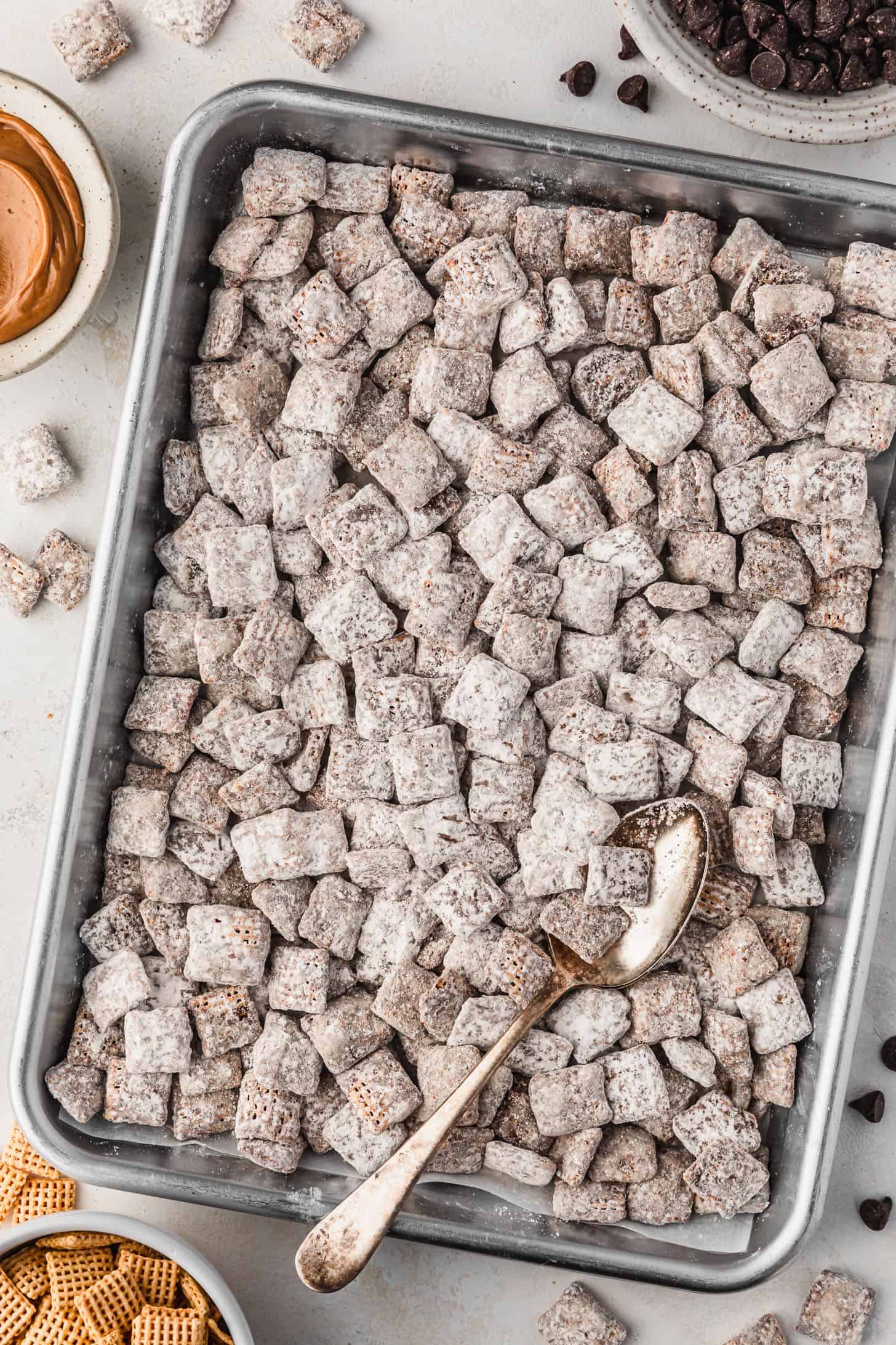 A silver pan of small batch puppy chow on a white counter next to white bowls of chocolate chips, peanut butter, and cereal.