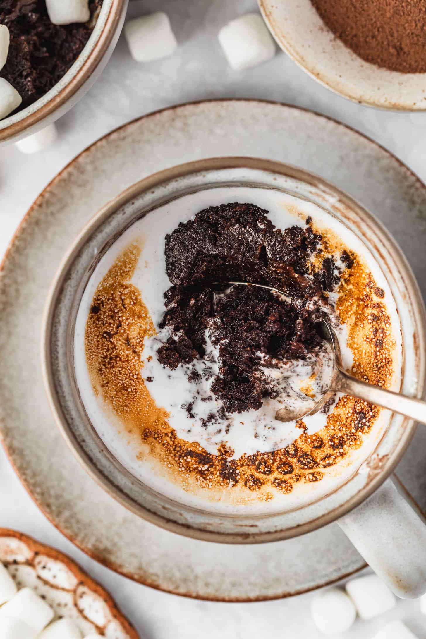 A grey mug with a hot chocolate mug cake on a white counter next to white bowls of hot cocoa mix and marshmallows.