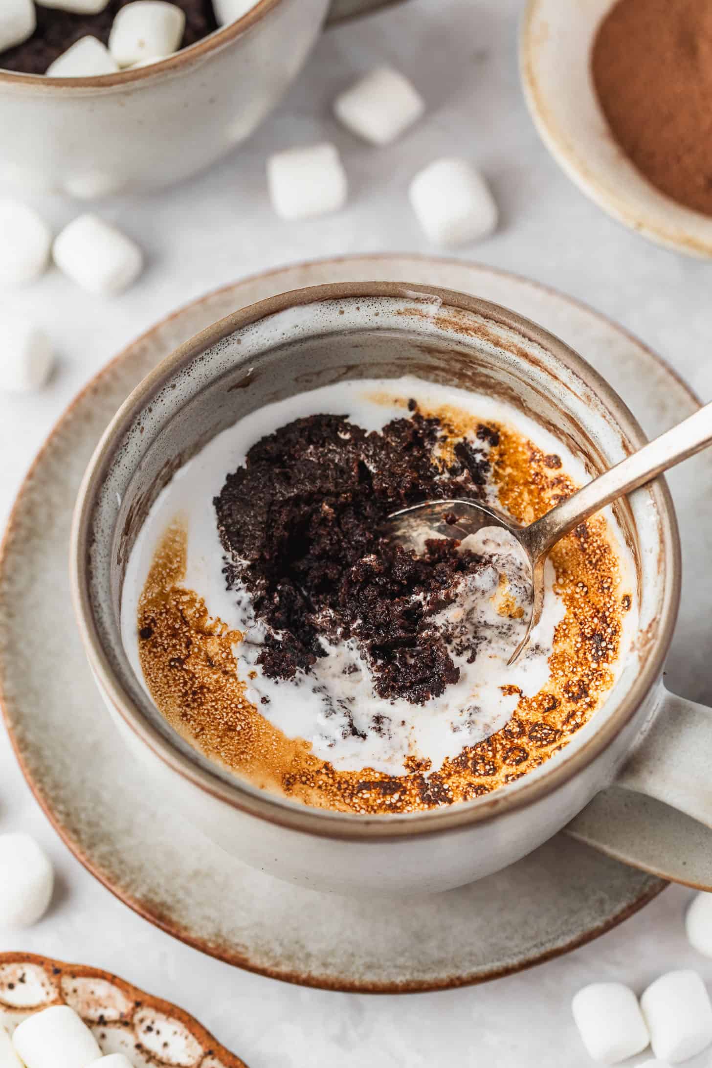 A grey mug with a hot chocolate mug cake and toasted marshmallow on a white marble table next to white bowls of marshmallows and cocoa mix.