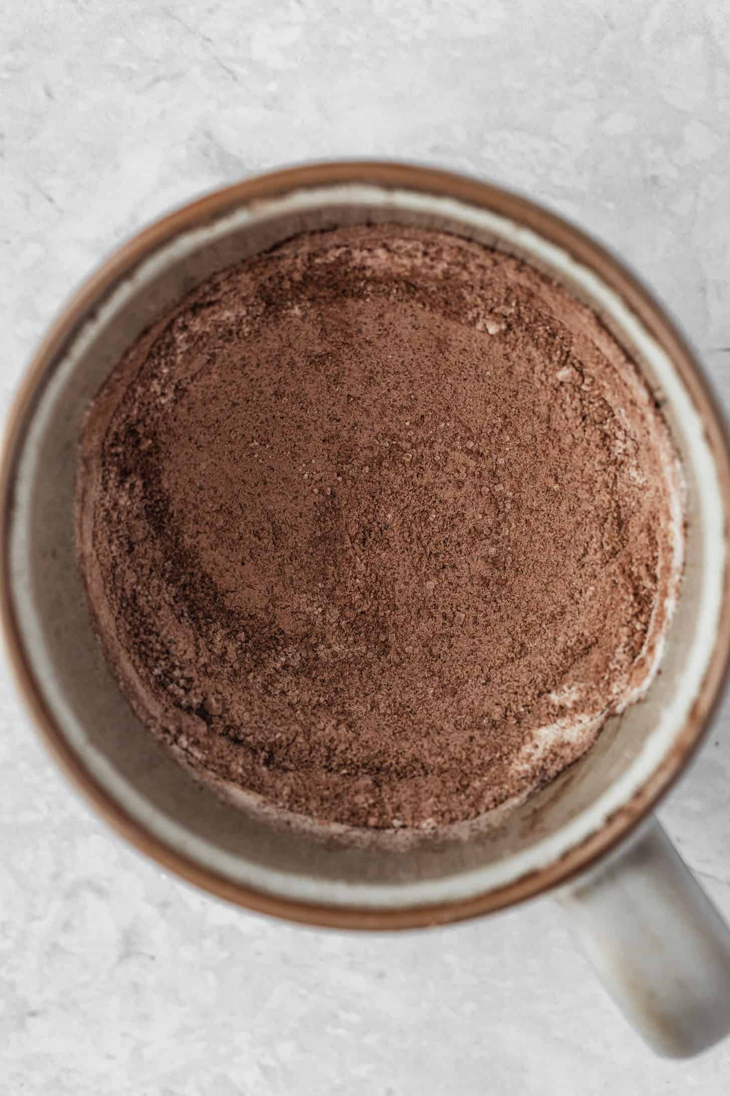 A grey mug with cocoa mix, sugar, and flour on a white counter.