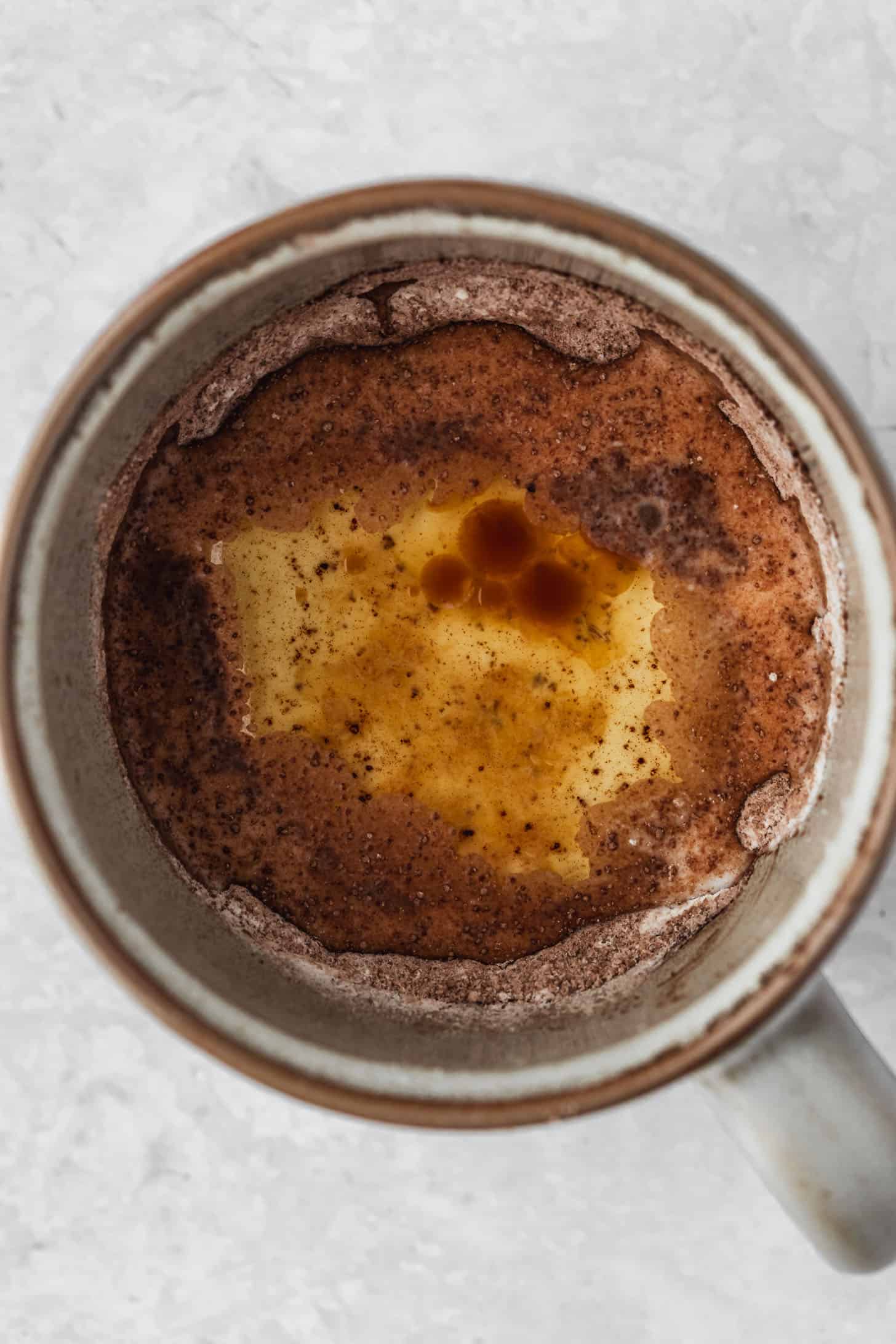A grey mug of cocoa mix, oil, milk, and vanilla on a white table.