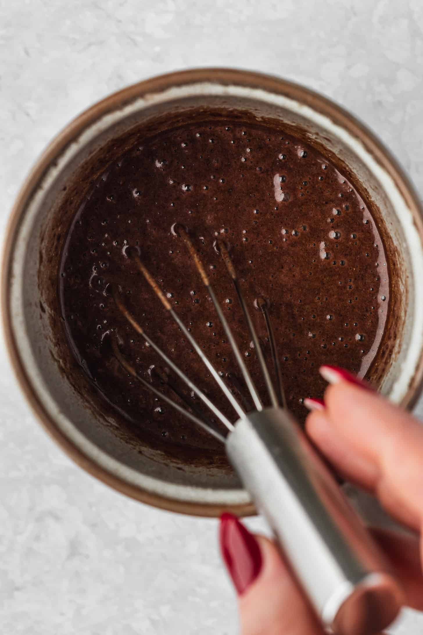 A hand using a whisk to mix chocolate batter in a grey mug.