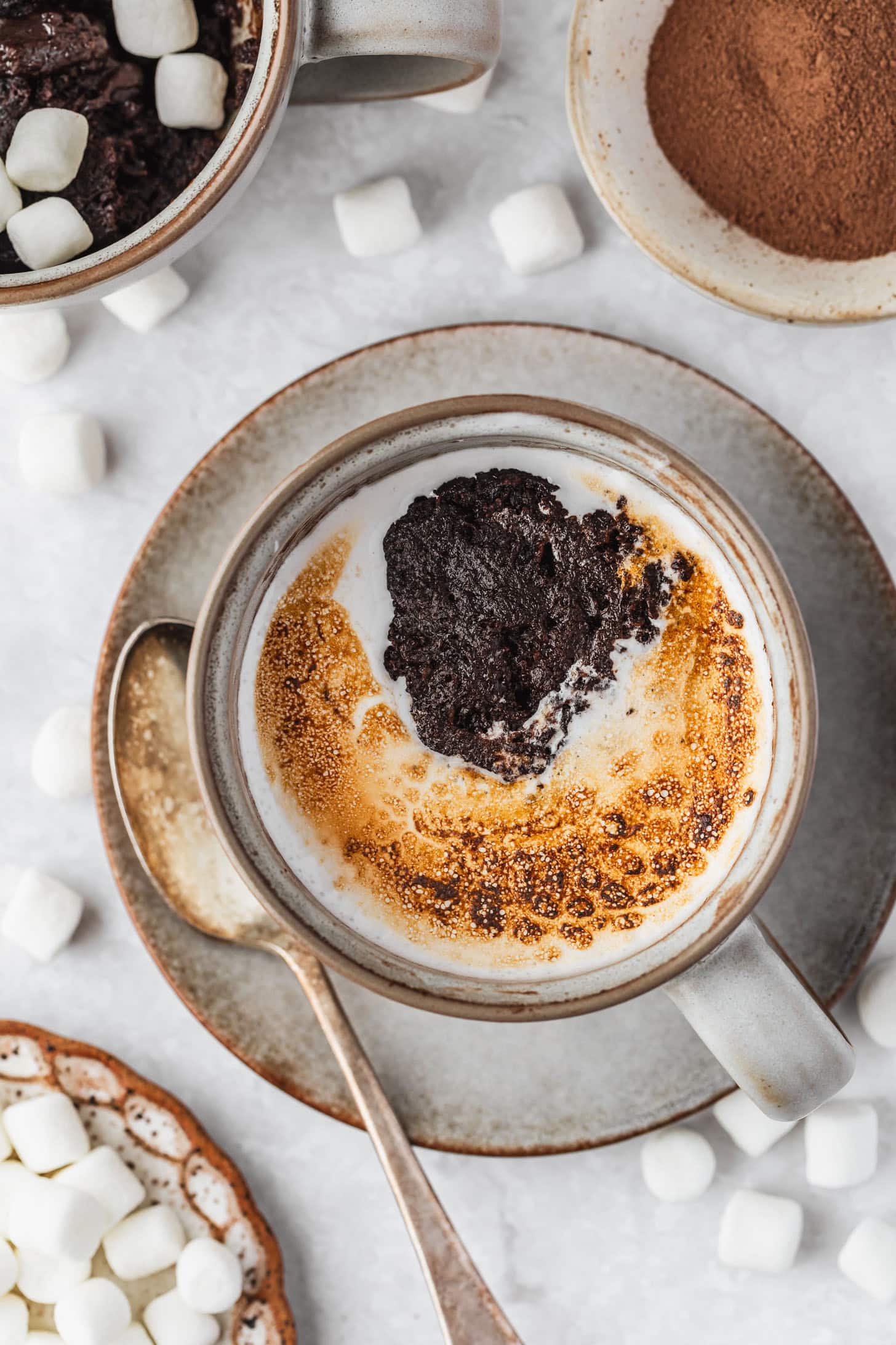 A hot cocoa mug cake on a white counter next to white bowls of hot chocolate mix and marshmallows.