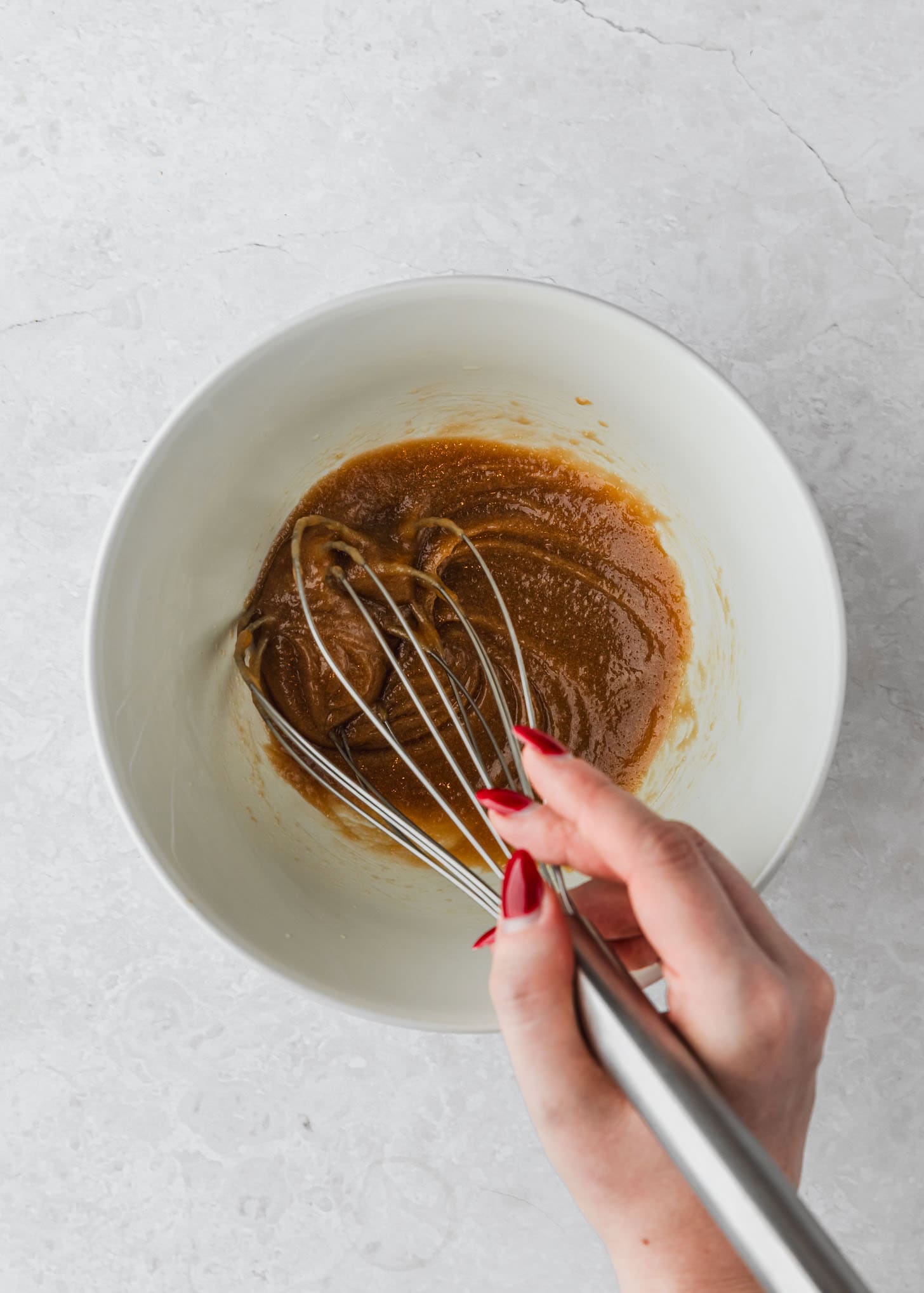 A hand using a whisk to mix butter and sugar in a white bowl.