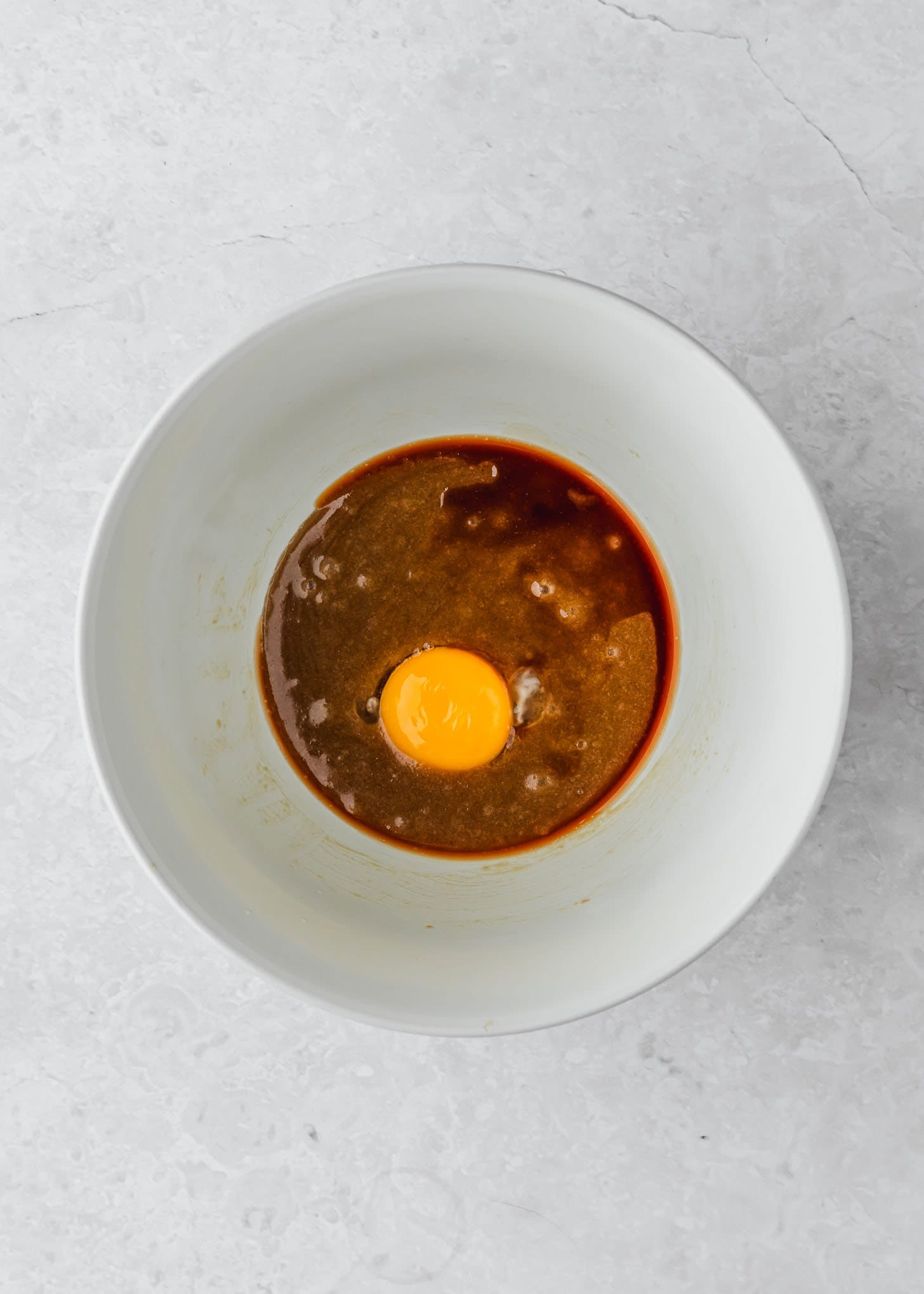 A white bowl of butter, sugar, vanilla, and an egg on a white table.