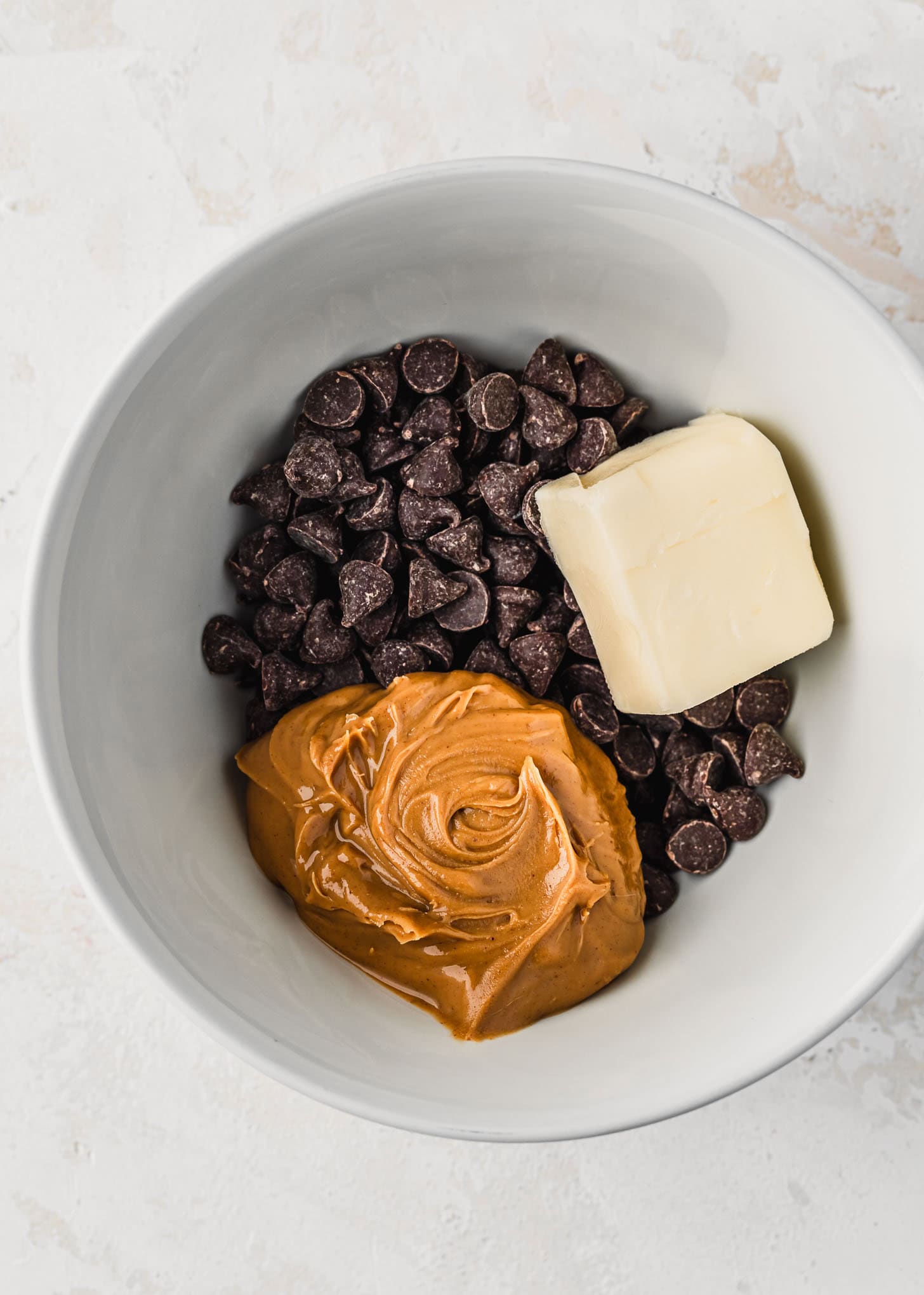 A white bowl of chocolate chips, peanut butter, and butter on a white table.