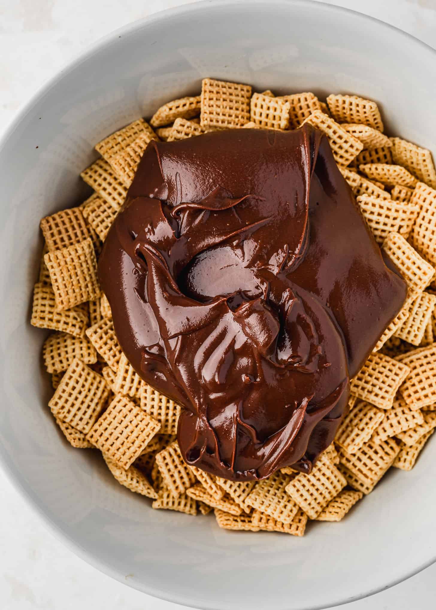 A white bowl of Chex and melted chocolate on a white counter.