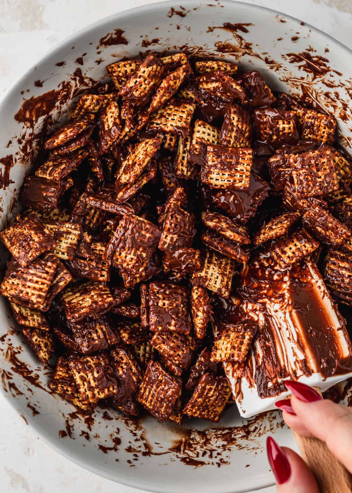 A hand using a rubber spatula to mix cereal and chocolate for puppy chow in a white bowl.