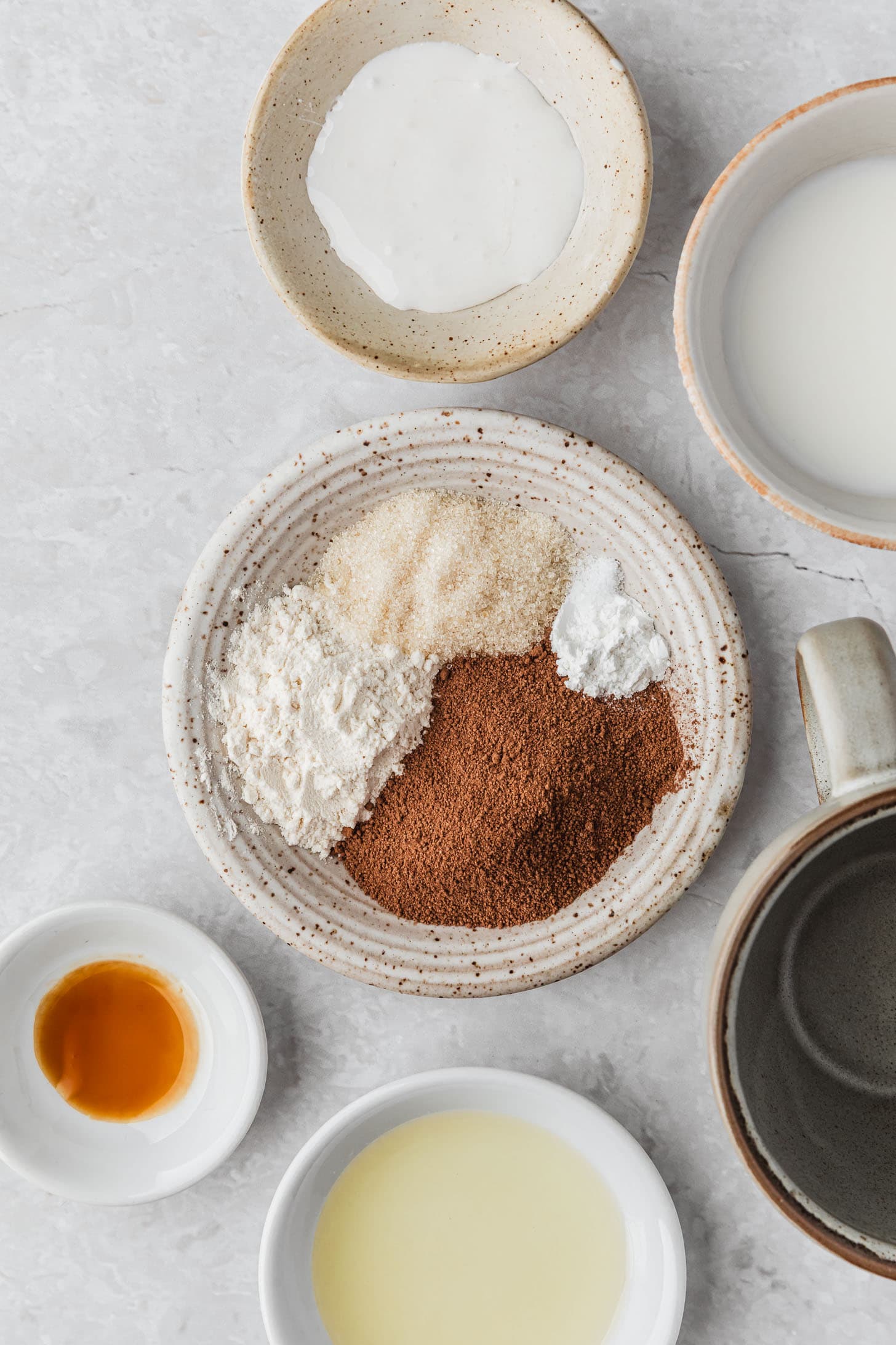 White bowls of cocoa mix, sugar, flour, milk, marshmallow creme, oil, and vanilla next to a grey mug on a white counter.
