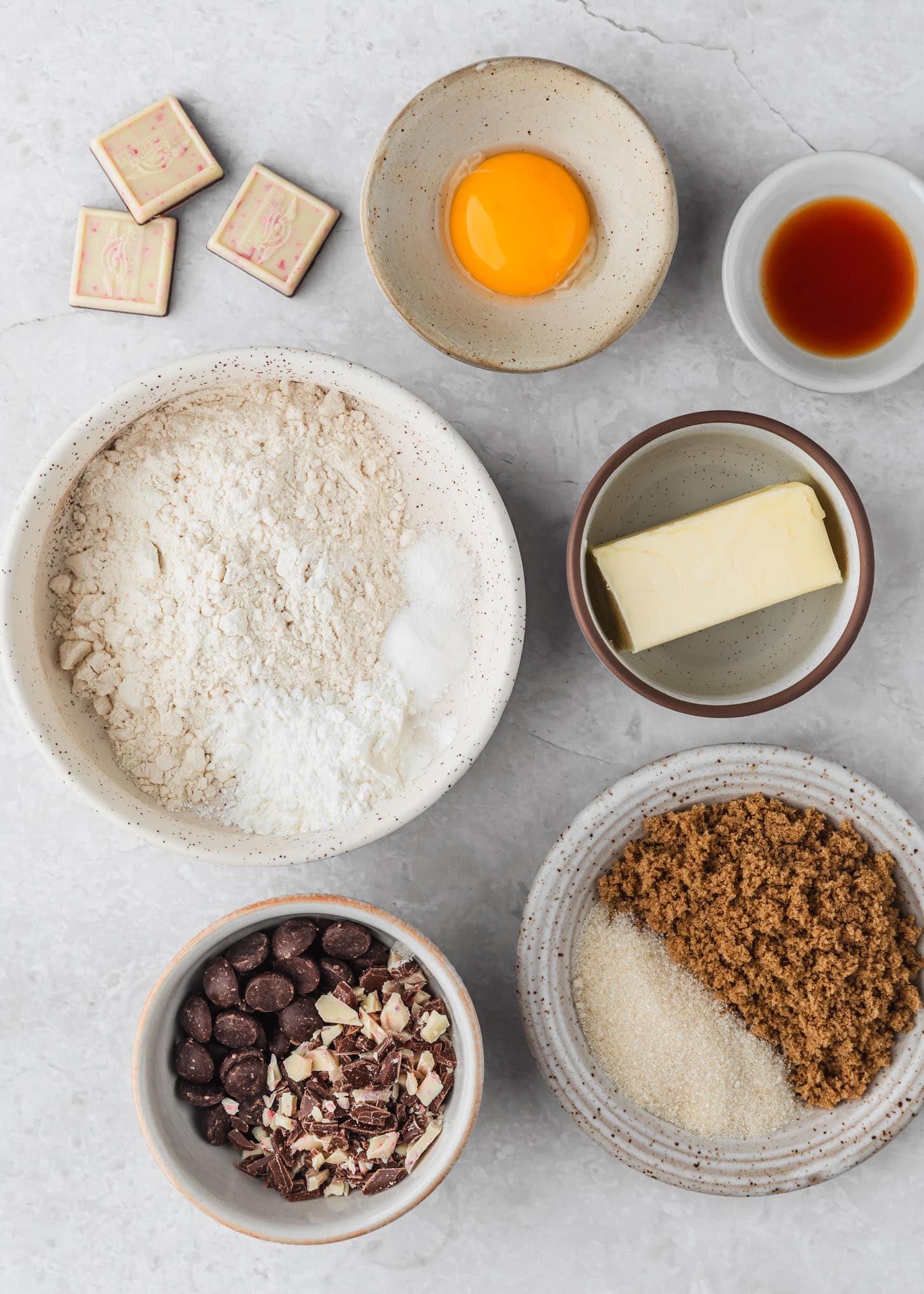 White and brown bowls of flour, brown sugar, white sugar, butter, vanilla, an egg, chocolate chips, and peppermint bark on a white counter.