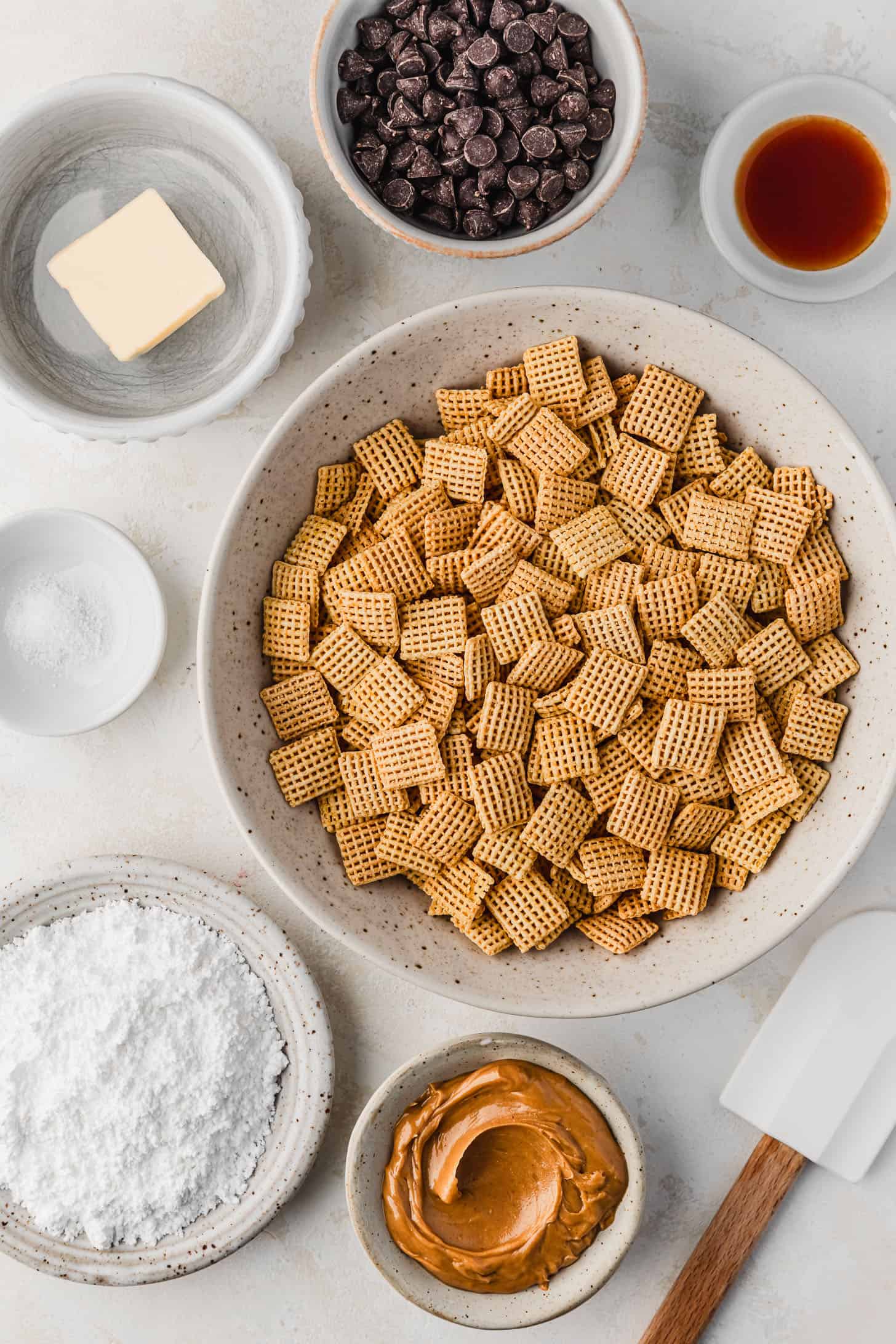 White bowls of Chex, chocolate, vanilla, butter, salt, powdered sugar, and peanut butter next to a white rubber spatula with a white background.