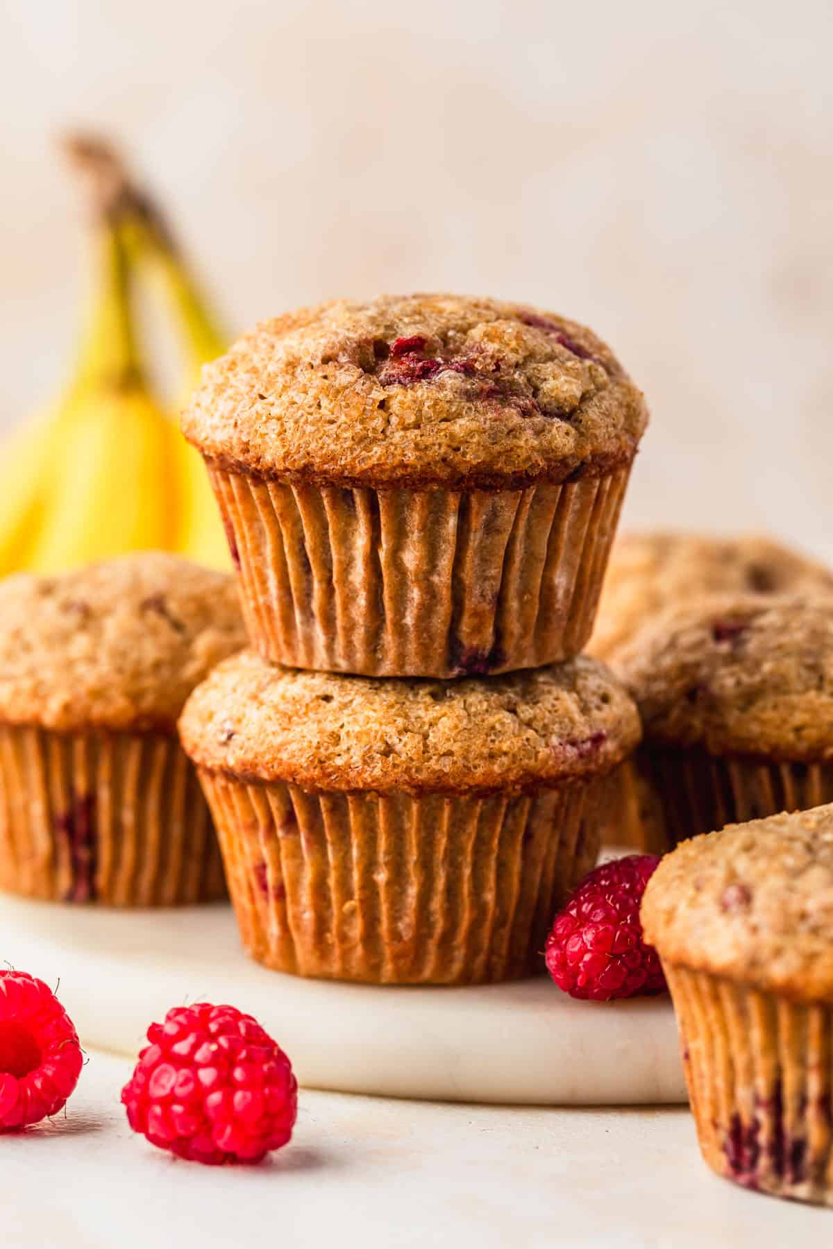 A stack of small batch raspberry banana muffins on a white tray next to more muffins, bananas, and raspberries on a tan background.