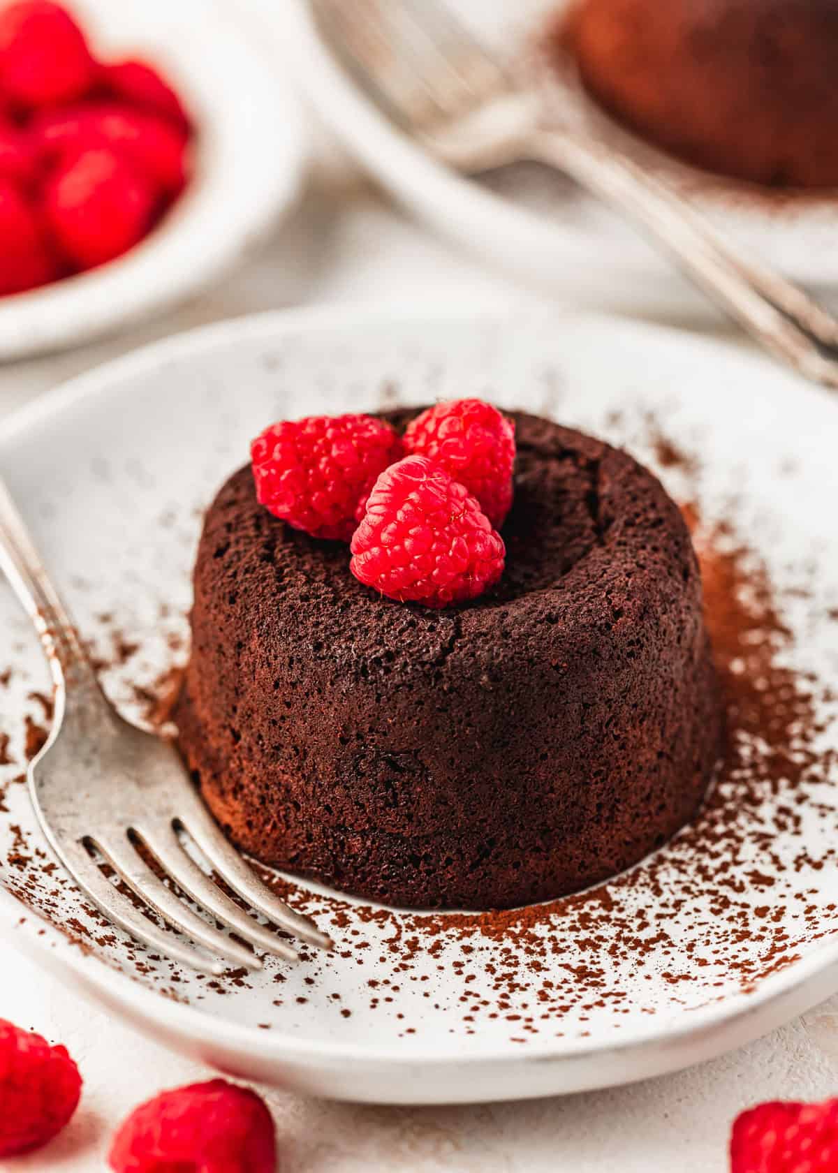 Two white plates with lava cakes for two, berries, and forks next to a white bowl of raspberries with a white background.