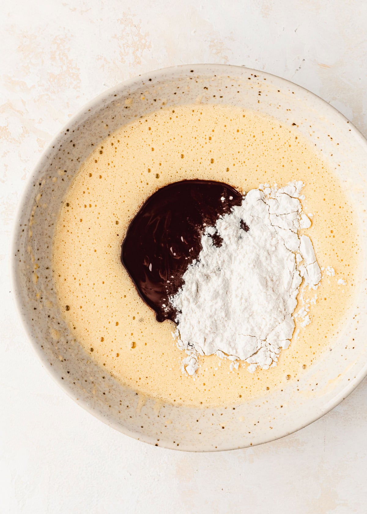 A white bowl of whisked eggs and sugar with flour and chocolate on a tan counter.