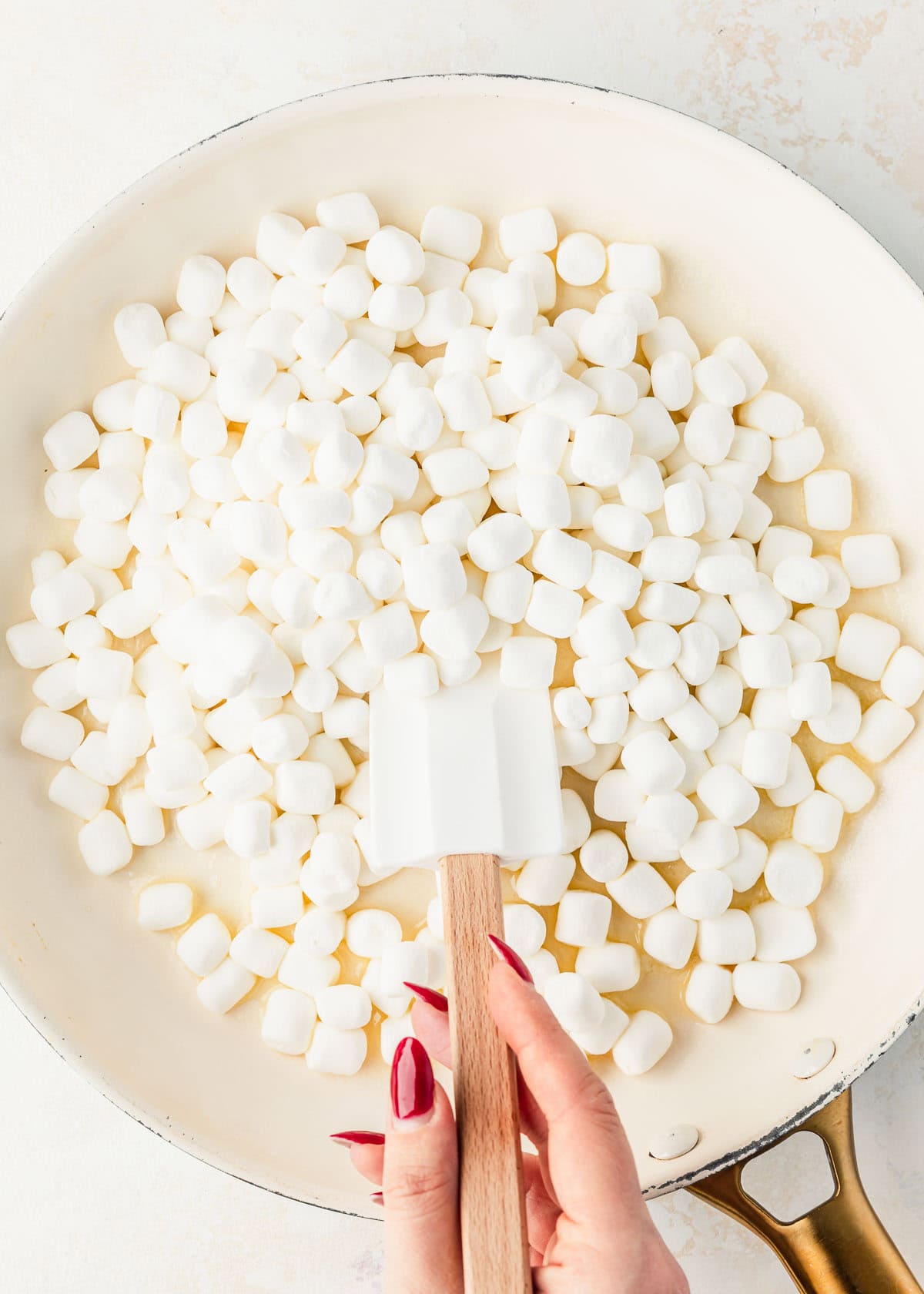 A hand using a spatula to stir marshmallows in a white pan.