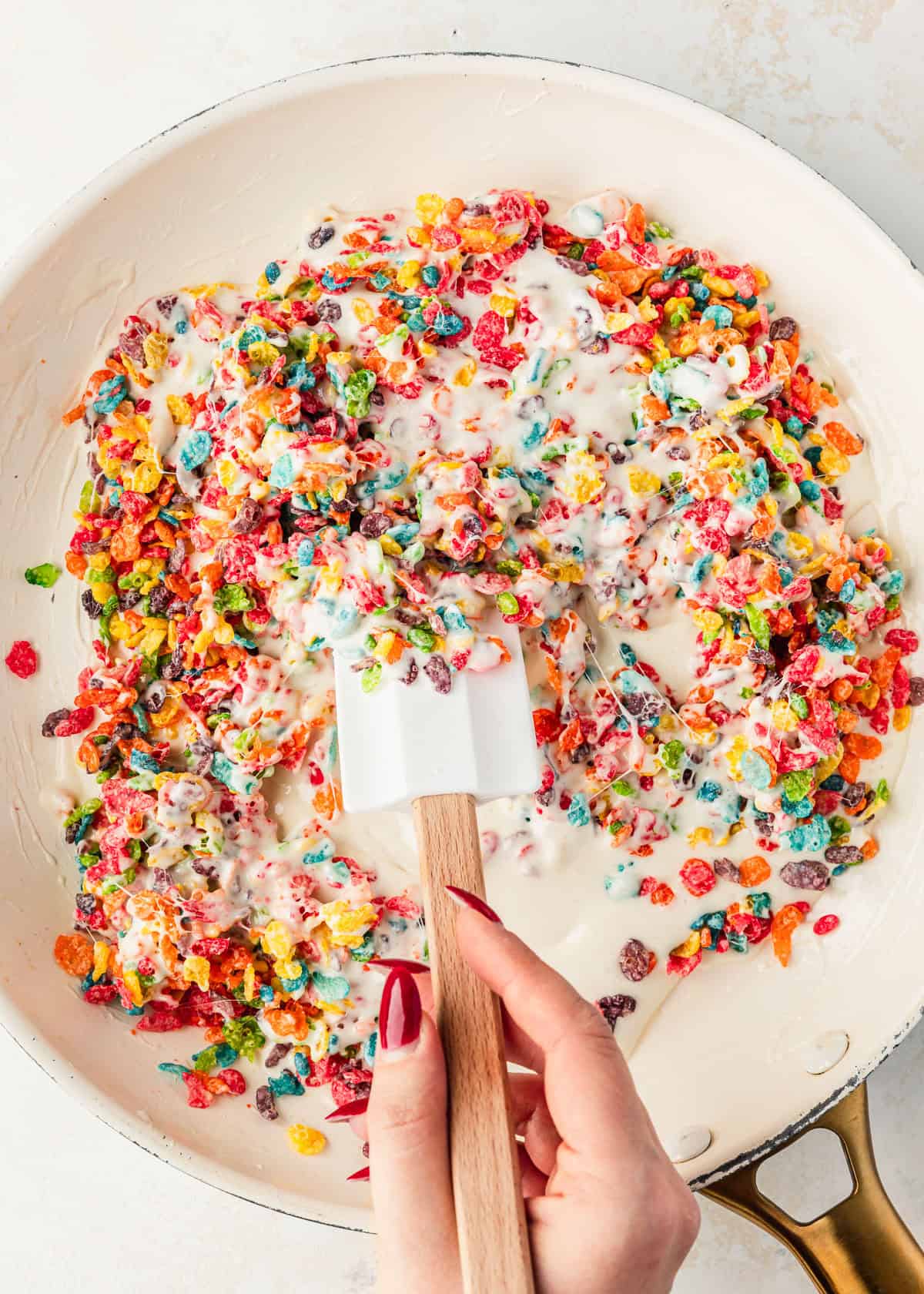 A hand using a rubber spatula to mix marshmallow Fruity Pebbles treats in a white pan.