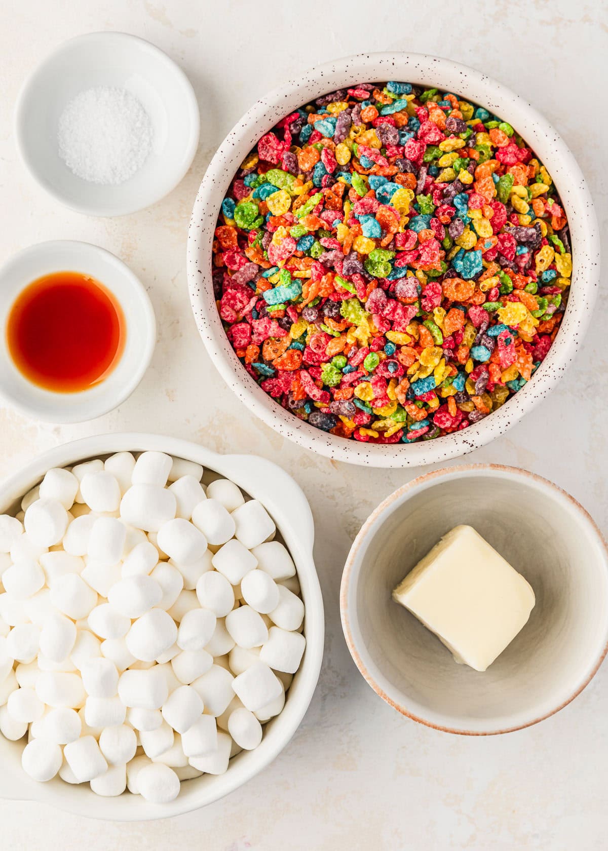 White bowls of marshmallows, fruit cereal, vanilla, salt, and butter on a beige counter.